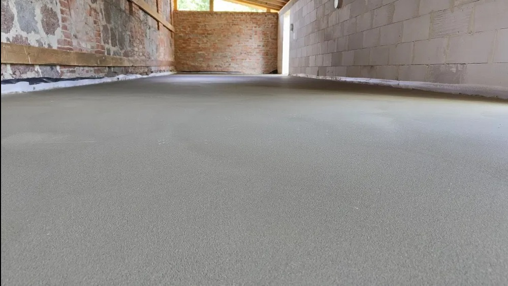 Gładka, szara posadzka jastrychowa w surowym wnętrzu z ceglanymi i betonowymi ścianami. Widoczny styk posadzki ze ścianą. Jasne światło dzienne.