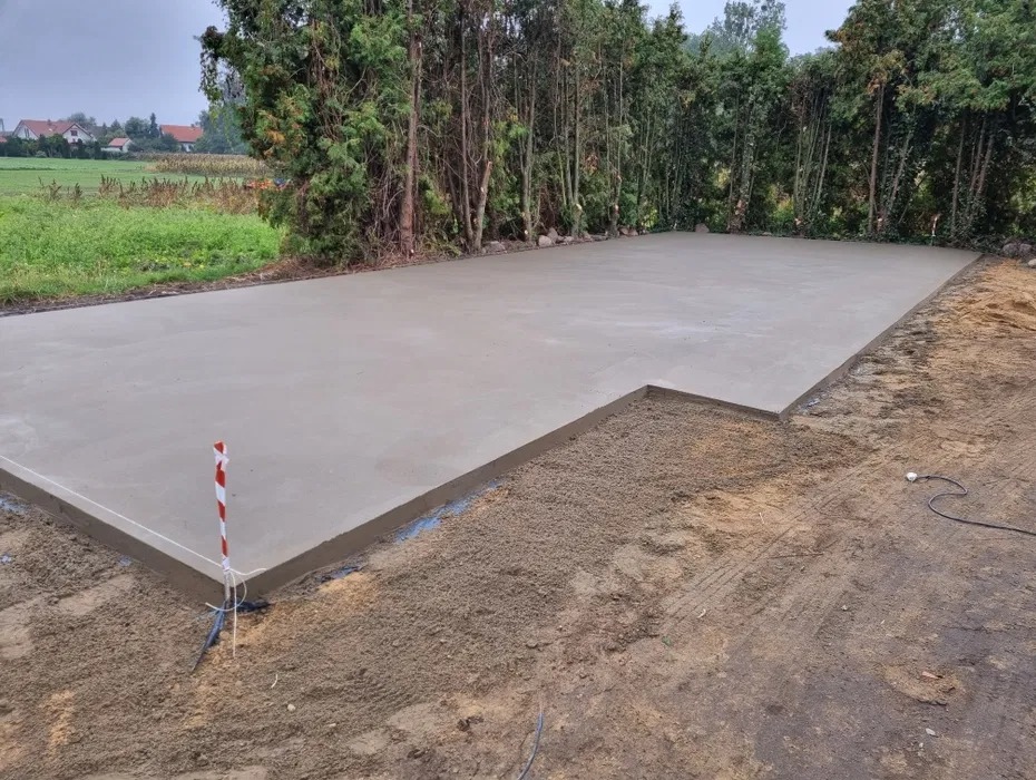 Świeżo wylana, gładka betonowa posadzka na zewnątrz, z widocznym obrysem i poziomowaniem, otoczona zielenią i drzewami. Widoczne ślady opon na ziemi.