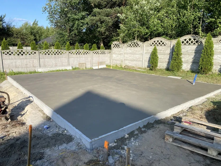 Świeżo wylany taras betonowy otoczony betonowym obrzeżem, z widocznym fragmentem ogrodzenia i zieleni w tle, w słoneczny dzień. Paleta i narzędzia obok.