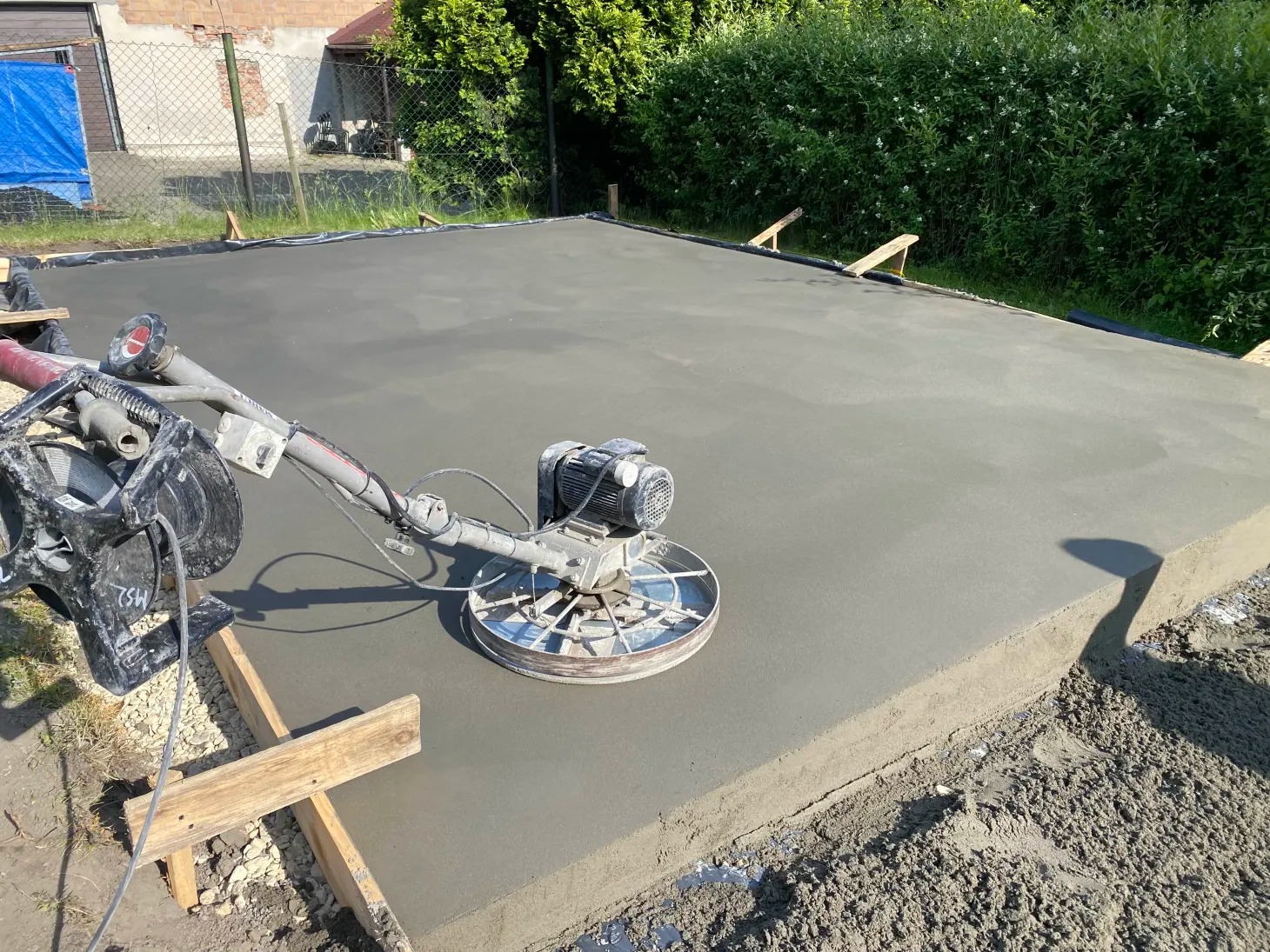 Wyrównywanie świeżo wylanej, szarej betonowej powierzchni w ogrodzie za pomocą zacieraczki mechanicznej. Widoczne drewniane szalunki.