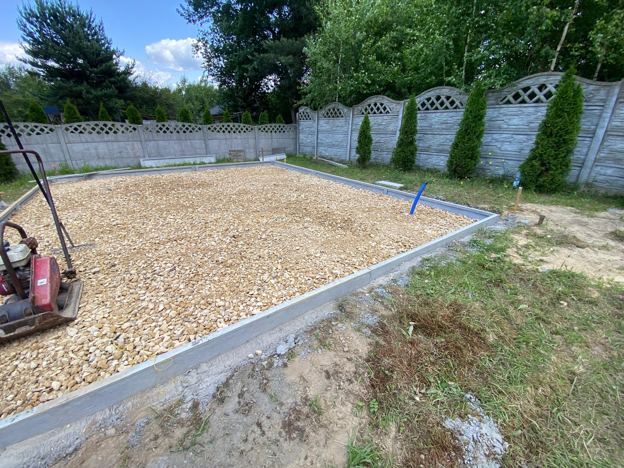 Przygotowanie podłoża pod taras: ubity żwir w betonowej ramie, zagęszczarka w rogu, ogrodzenie z prefabrykatów betonowych w tle, zielone tuje.