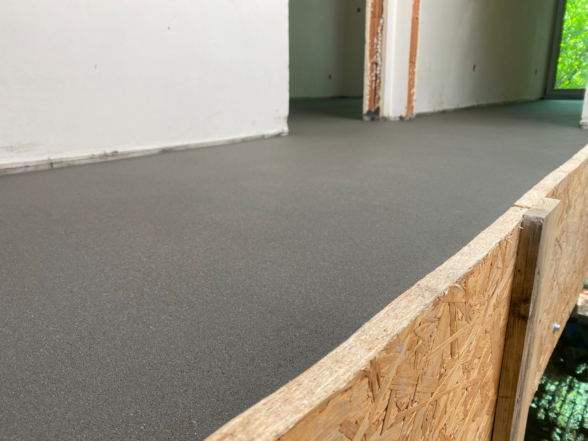 Świeżo wylana, gładka posadzka jastrychowa z widocznym deskowaniem, surowy stan wykończenia wnętrza, naturalne światło z okna w tle.