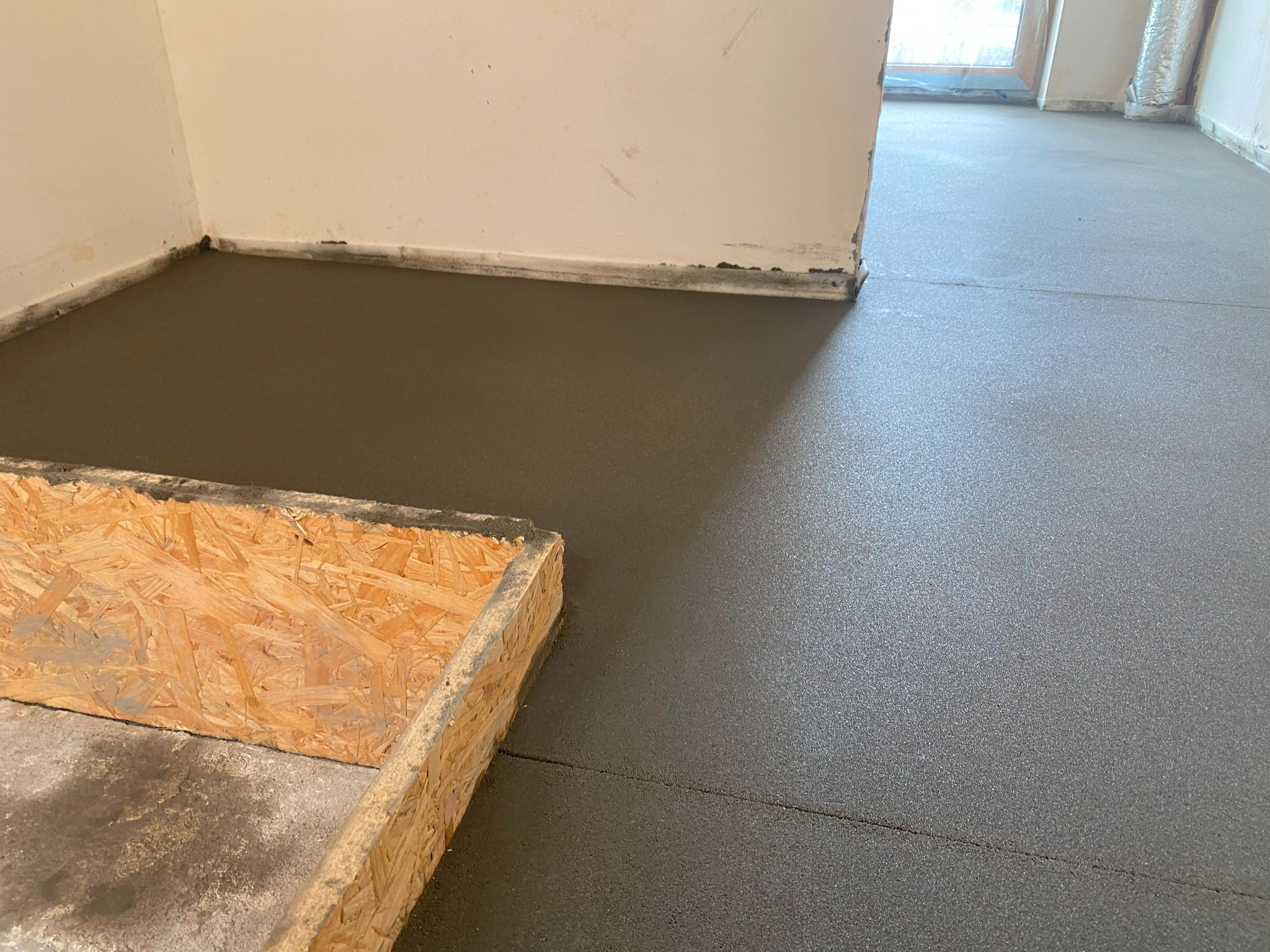 Świeżo wylana, gładka posadzka jastrychowa w jasnym pomieszczeniu, widoczna drewniana forma w rogu. Ślady po listwach przypodłogowych przy ścianach.