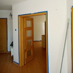 IURII PYNKO - Przygotowanie do malowania: ściany i futryny zabezpieczone taśmą malarską, widoczny wałek oparty o ścianę, drewniana podłoga. W tle pomalowane drzwi.