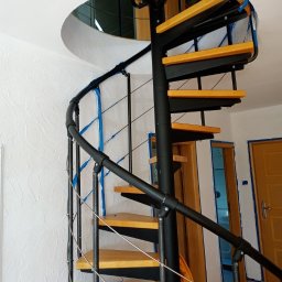 IURII PYNKO - Spiralne schody z drewnianymi stopniami i czarną metalową konstrukcją, zabezpieczone taśmą malarską podczas remontu, widok na ścianę z fakturą.