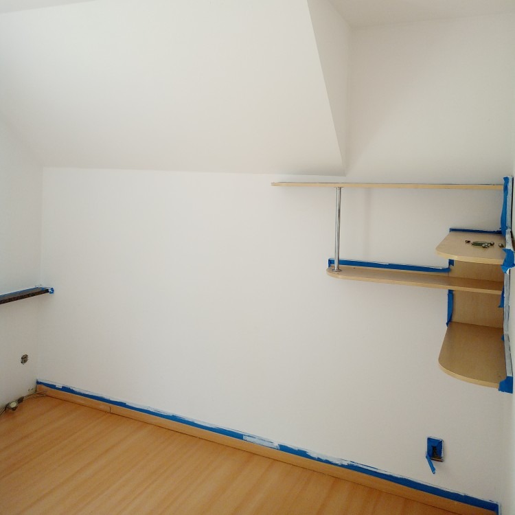 Białe wnętrze pokoju na poddaszu, przygotowane do malowania. Widoczne zabezpieczenia taśmą malarską przy podłodze i półkach. Świeżo pomalowane ściany.