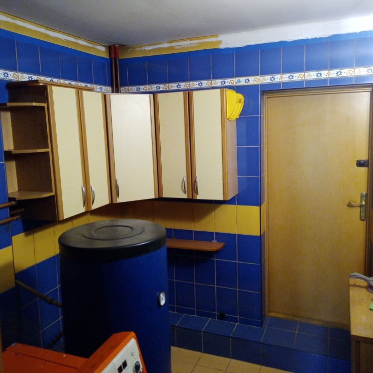 Pomieszczenie z niebiesko-żółtymi kafelkami, kremowymi szafkami i bojlerem. Świeżo pomalowany sufit, widoczne niedociągnięcia. Żółta szpachelka na ścianie.