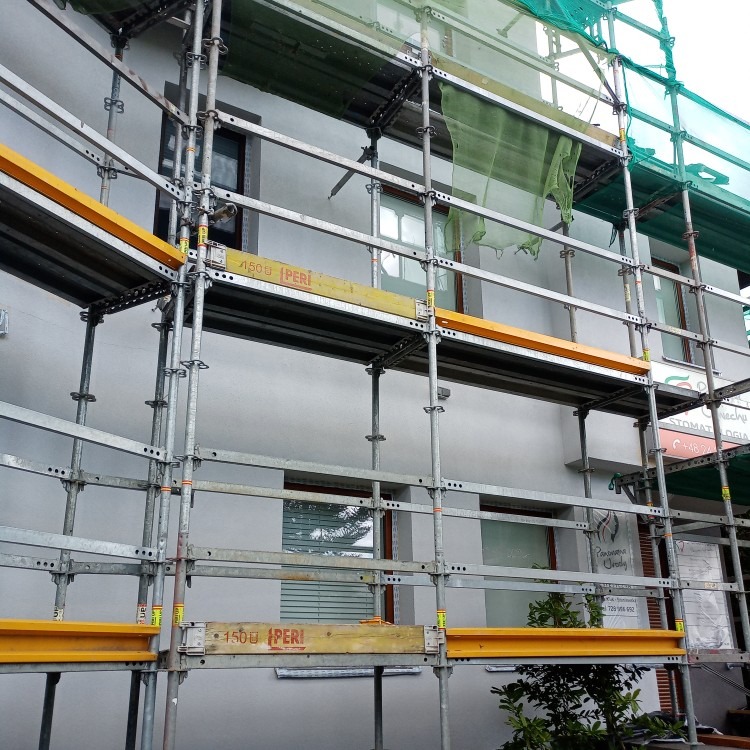 Elewacja budynku w szarościach, z widocznymi rusztowaniami i siatką ochronną, sugerująca prace remontowe lub malarskie w toku.