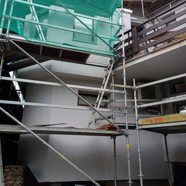 Biała elewacja domu w trakcie prac wykończeniowych, widoczna z poziomu rusztowania z siatką ochronną. Okna z brązowymi ramami.