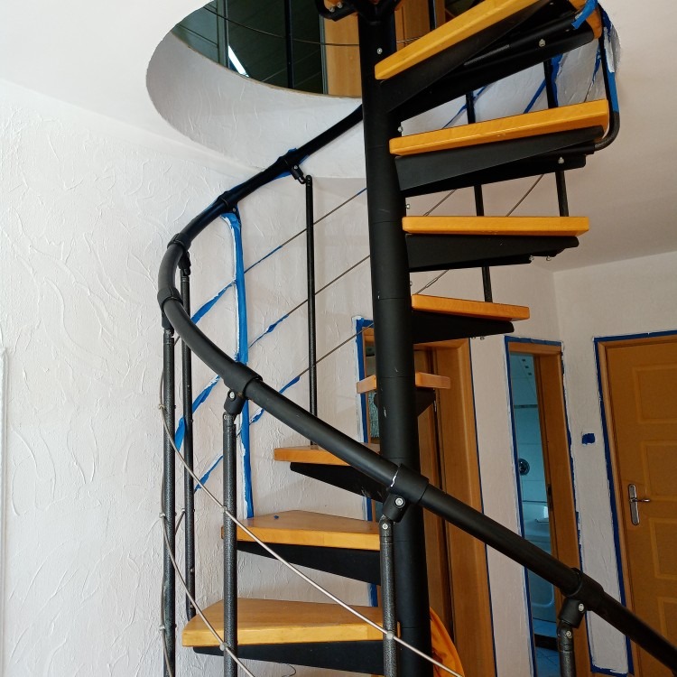 Spiralne schody z drewnianymi stopniami i czarną metalową konstrukcją, zabezpieczone taśmą malarską podczas remontu, widok na ścianę z fakturą.