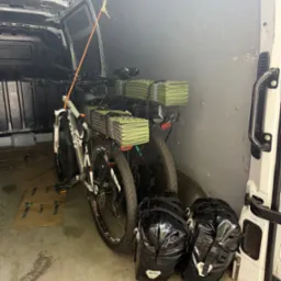 Wnętrze busa z dwoma rowerami górskimi i bagażami, zabezpieczonymi pasami transportowymi. Widoczne kartony na podłodze i metalowe ściany pojazdu.