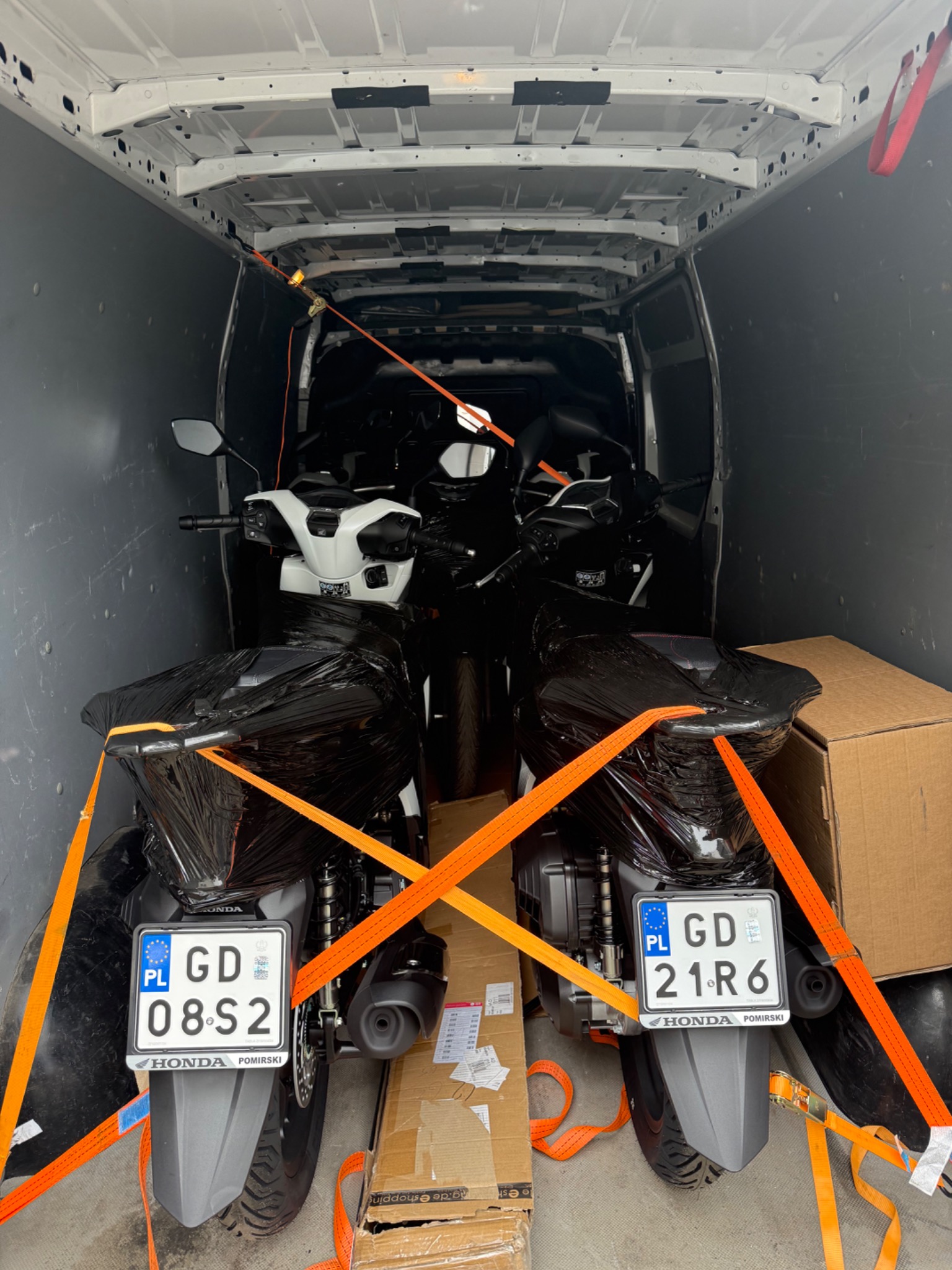 Wnętrze busa z zabezpieczonymi motocyklami Honda, owiniętymi folią i spiętymi pomarańczowymi pasami transportowymi. Widoczne tablice rejestracyjne.