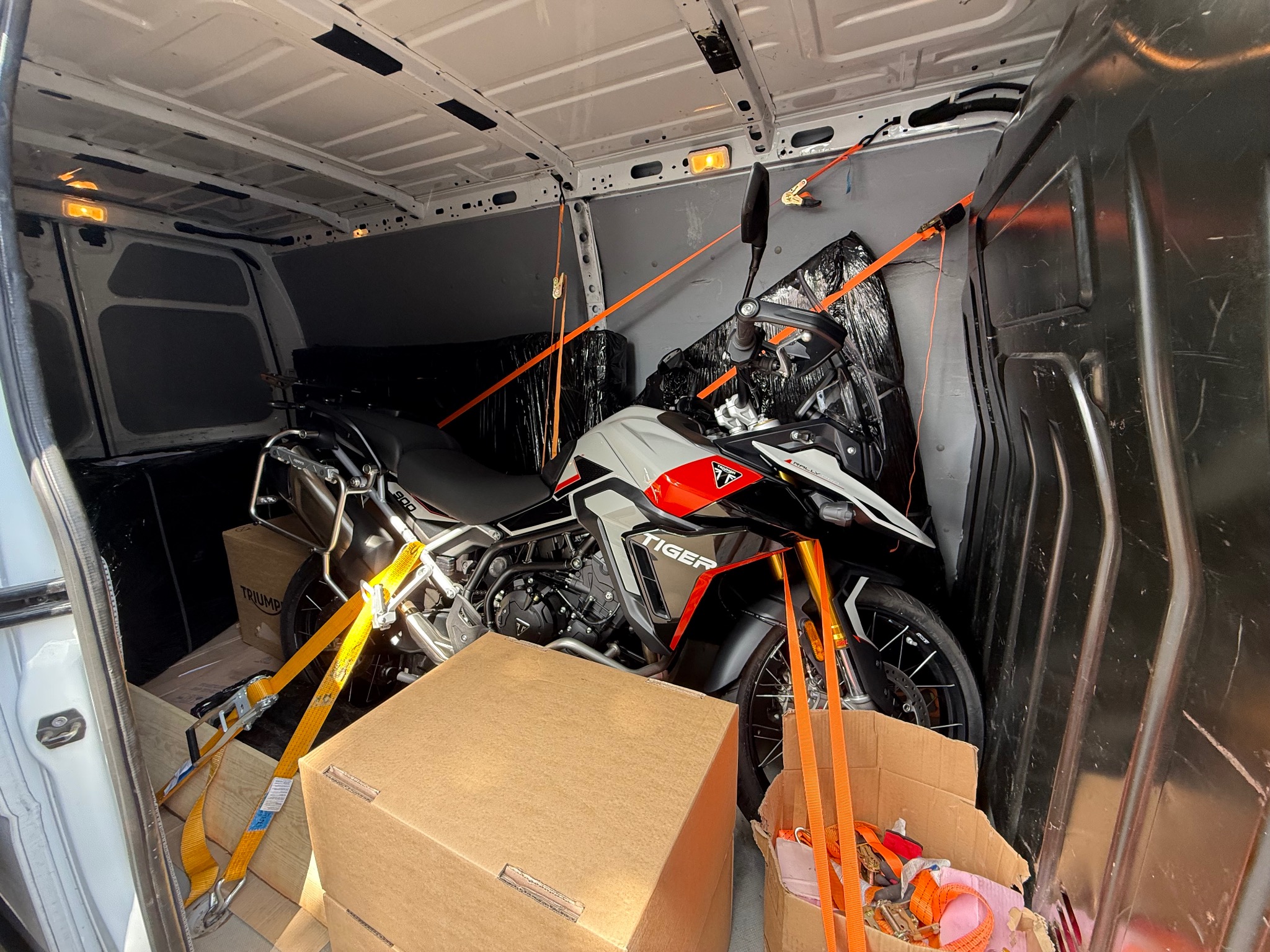 Motocykl Triumph Tiger 900 zabezpieczony pasami wewnątrz białego busa, gotowy do transportu. Kartony obok motocykla. Widok z wnętrza pojazdu.