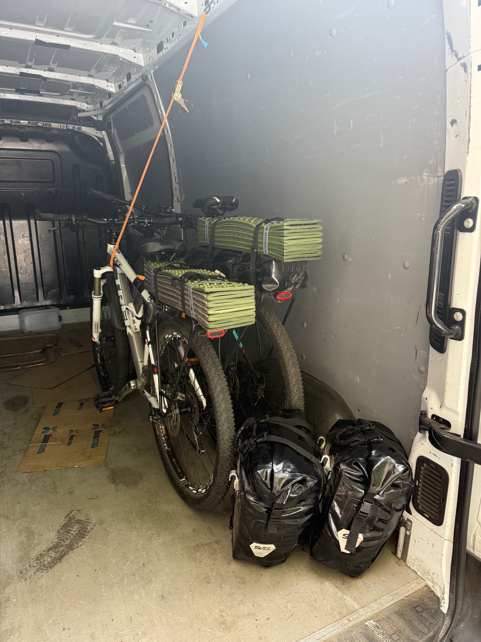 Wnętrze busa z dwoma rowerami górskimi i bagażami, zabezpieczonymi pasami transportowymi. Widoczne kartony na podłodze i metalowe ściany pojazdu.