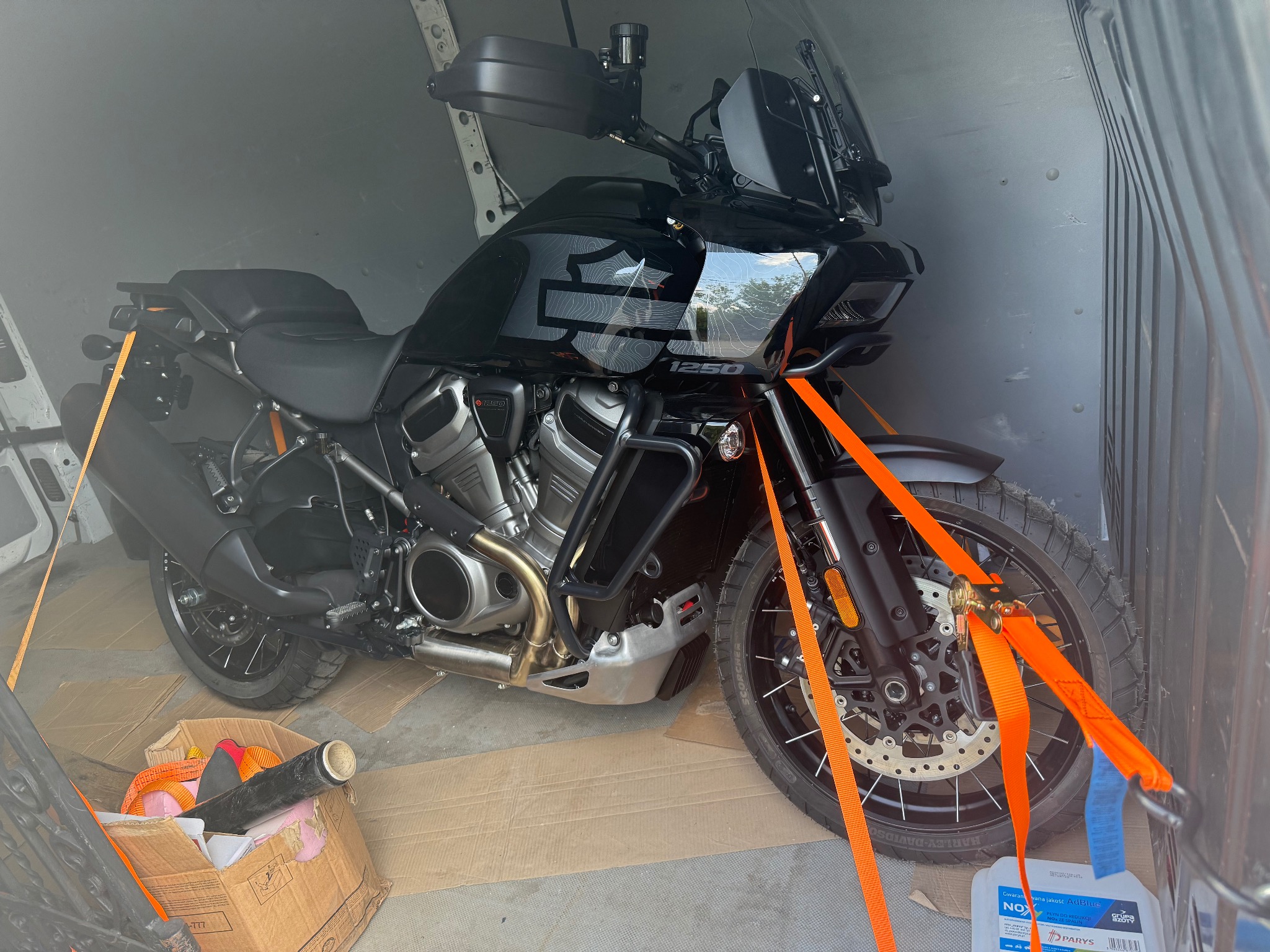 Czarny motocykl Harley-Davidson 1250, zabezpieczony pomarańczowymi pasami transportowymi wewnątrz szarej furgonetki, gotowy do przewozu. Widoczny karton i płyn AdBlue.