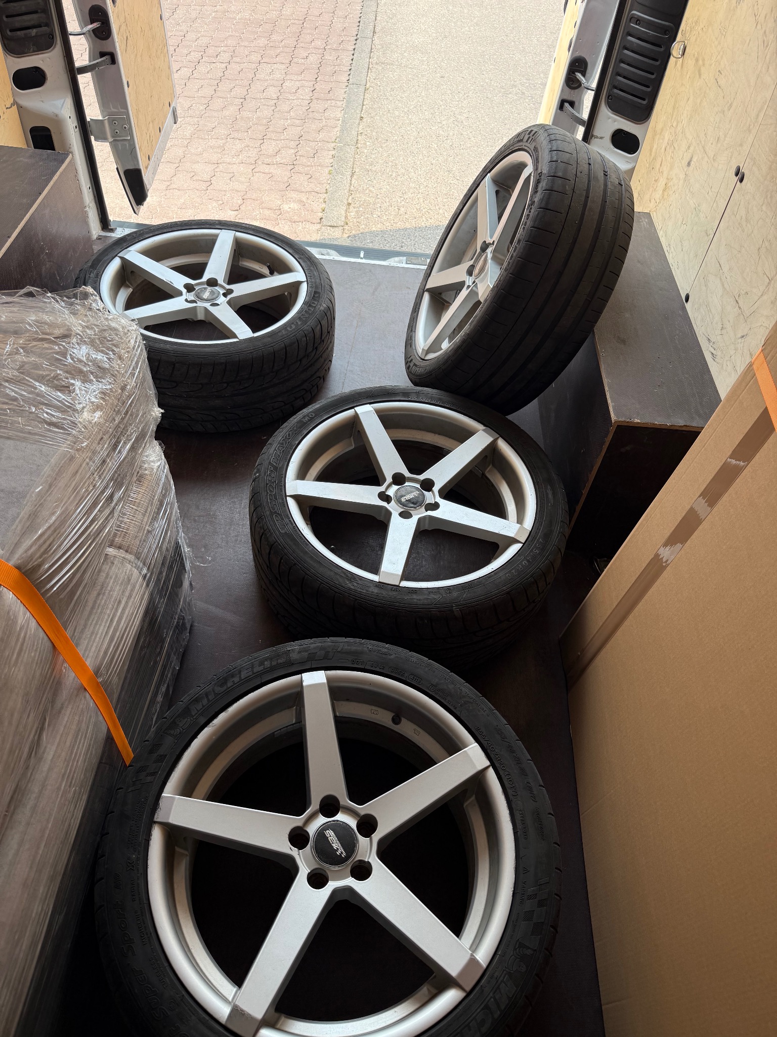 Cztery aluminiowe felgi z oponami w przestrzeni ładunkowej busa, obok zapakowane kartony i folia stretch. Widok z góry, lekki kąt.