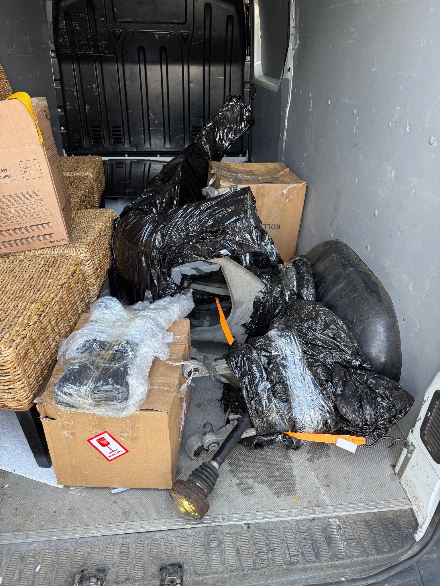 Wnętrze busa załadowane kartonami, wiklinowymi koszami i elementami samochodowymi owiniętymi w czarne worki i folię. Widoczny znak 'Uwaga szkło'.