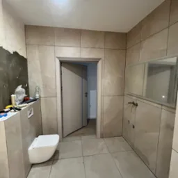 Łazienka w trakcie wykańczania: beżowe płytki imitujące marmur na ścianach i podłodze, wisząca toaleta, umywalka i lustro. Widoczne narzędzia i materiały budowlane.