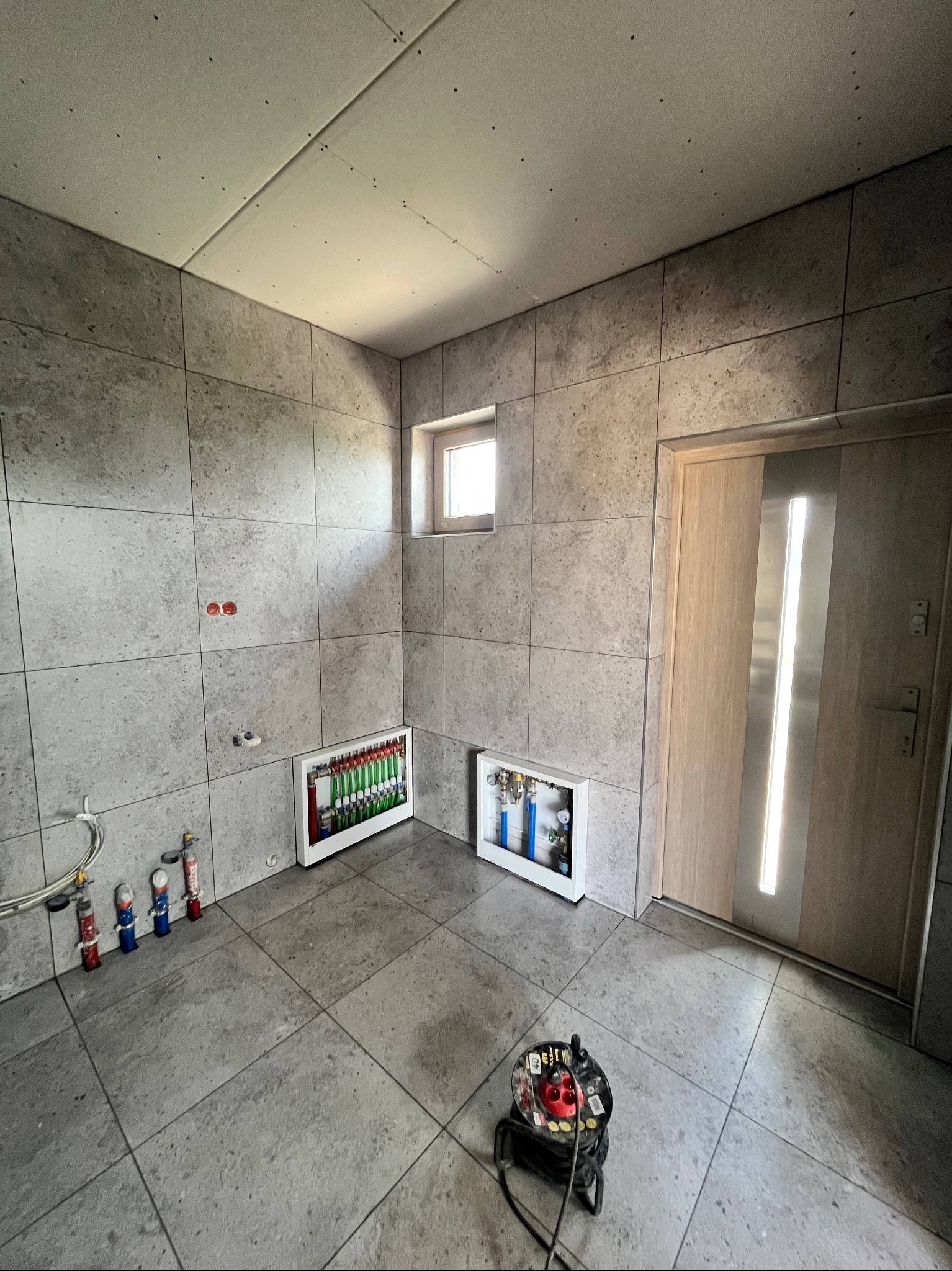 Łazienka w trakcie prac wykończeniowych: szare płytki na ścianach i podłodze, widoczne instalacje wodne i elektryczne, narzędzia. Małe okno, drzwi z przeszkleniem.