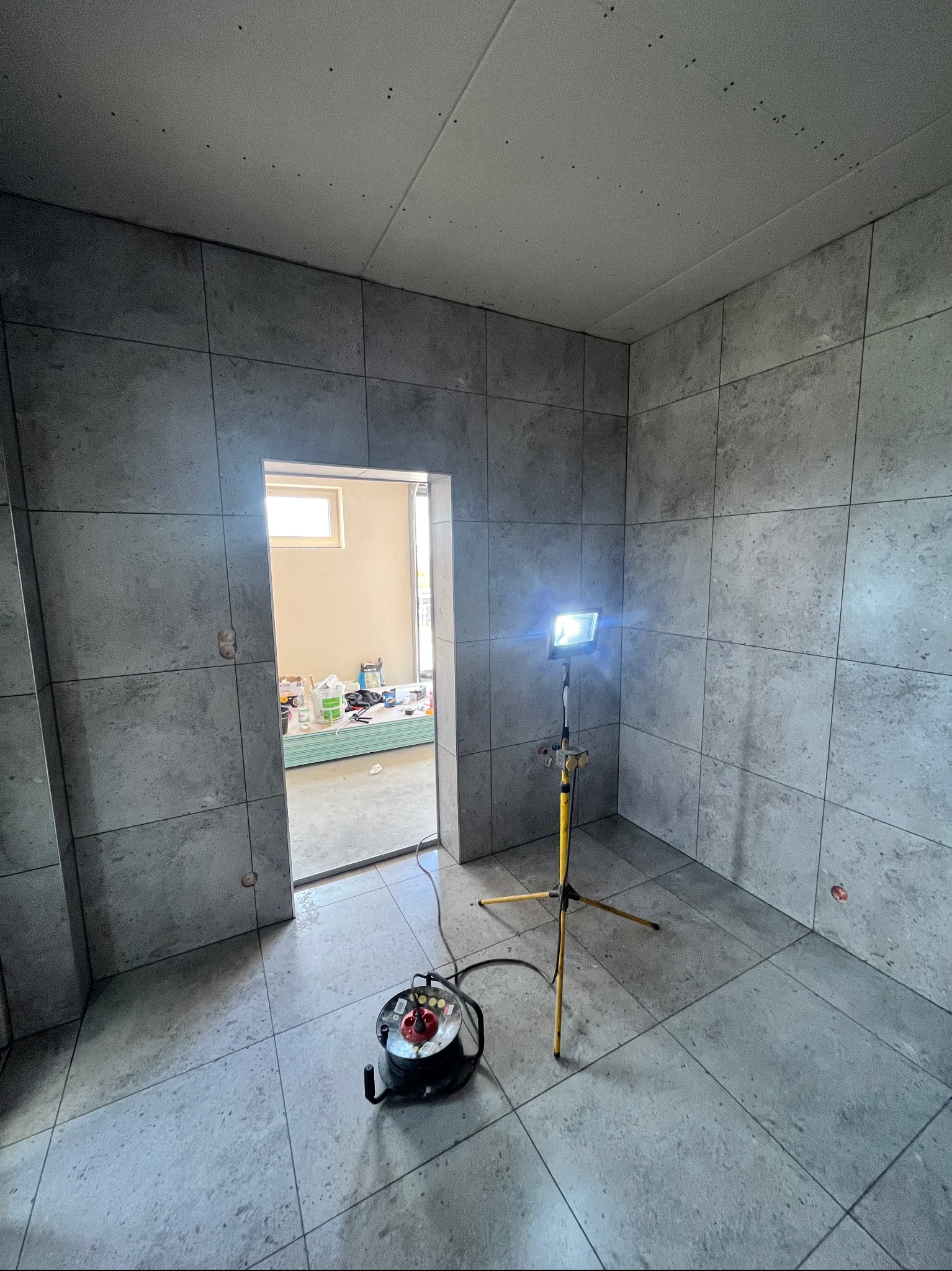 Wnętrze łazienki w trakcie wykańczania, z szarymi płytkami na ścianach i podłodze. Widoczny halogen na statywie i otwarte drzwi do innego pomieszczenia.