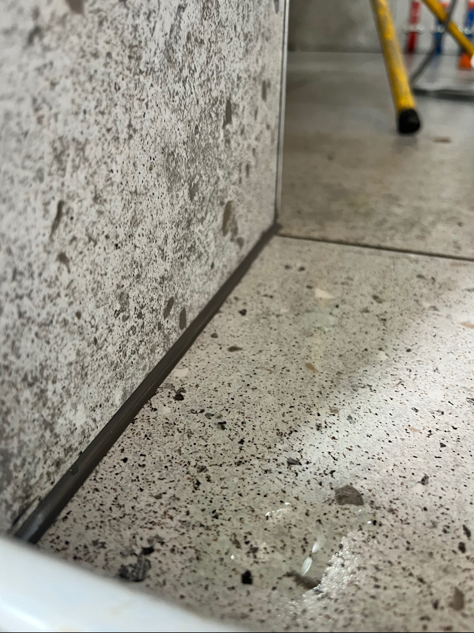 Połączenie ściany i podłogi wykończone szarą fugą. Płytki imitujące kamień o porowatej strukturze. Widoczne poziomice w tle.