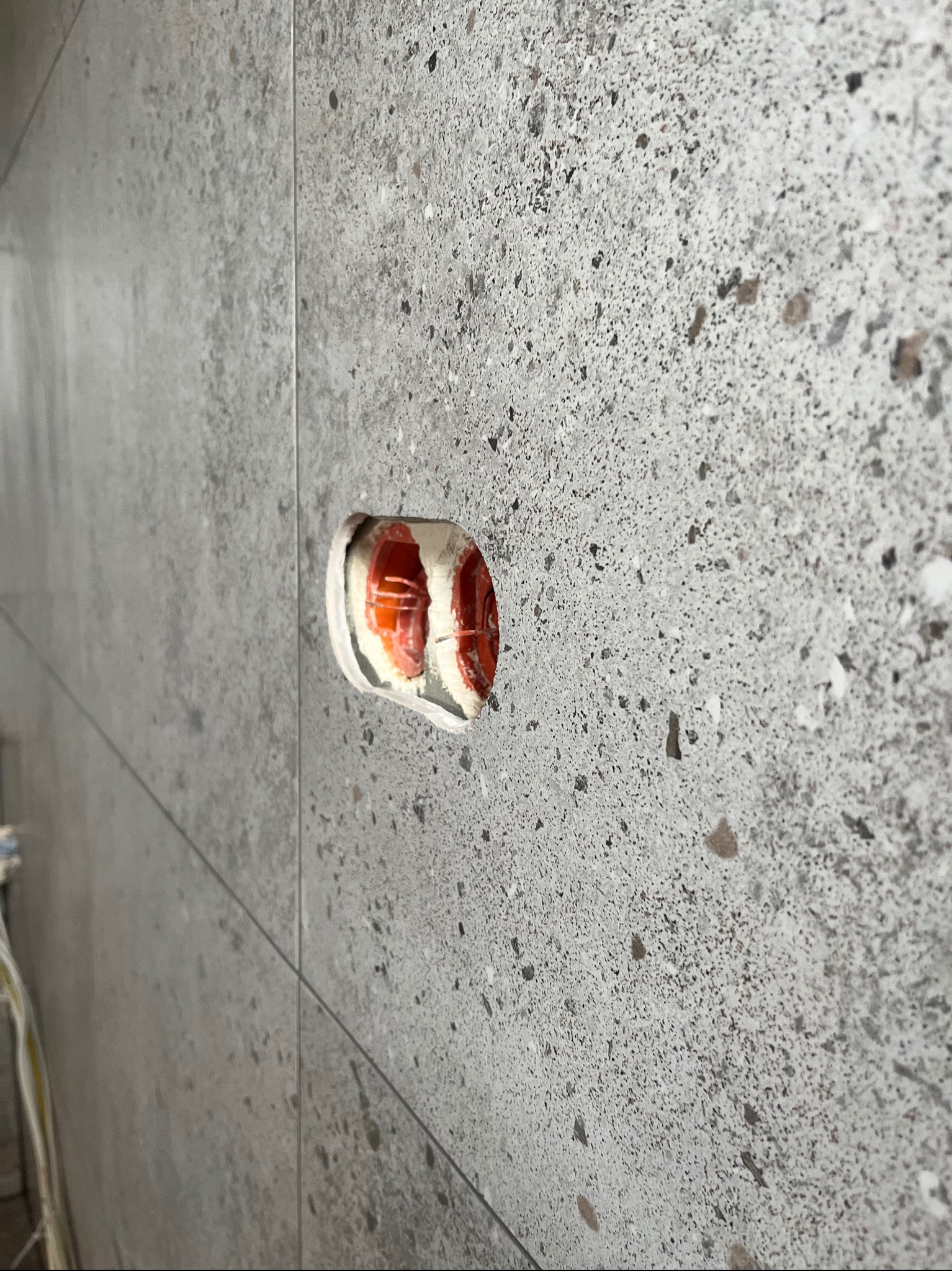 Szara, matowa płytka ścienna z widocznym otworem pod puszkę elektryczną, z widocznymi kablami w środku. Widok z boku, lekko z dołu.