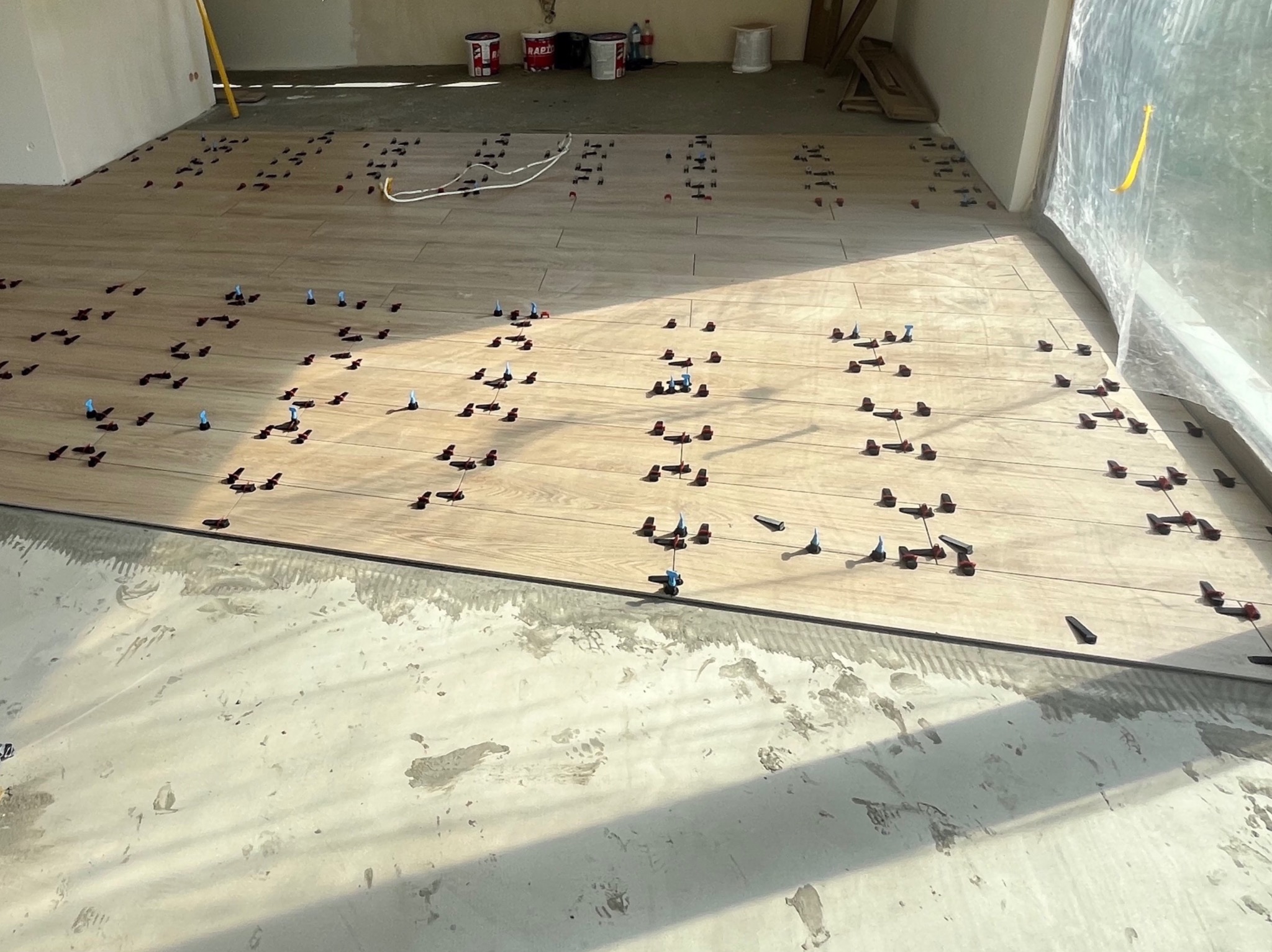 Układanie płytek podłogowych w jasnym odcieniu drewna, z użyciem klipsów poziomujących. Widoczne wiaderka z zaprawą w tle, częściowo zakryte folią okno.