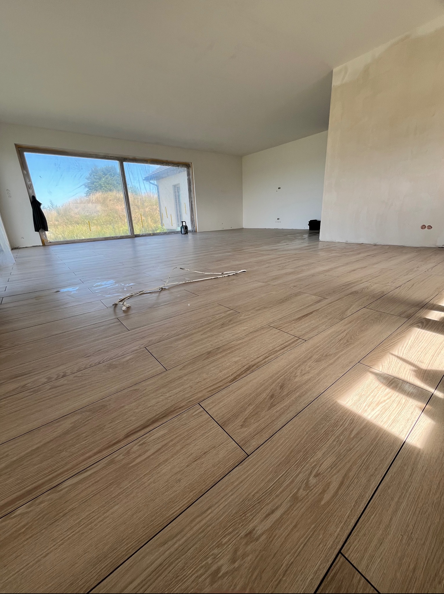 Jasne płytki podłogowe imitujące drewno, ułożone w salonie z dużym oknem. Świeżo położona podłoga w nowym domu. Na ścianach widoczne ślady po gładzi.
