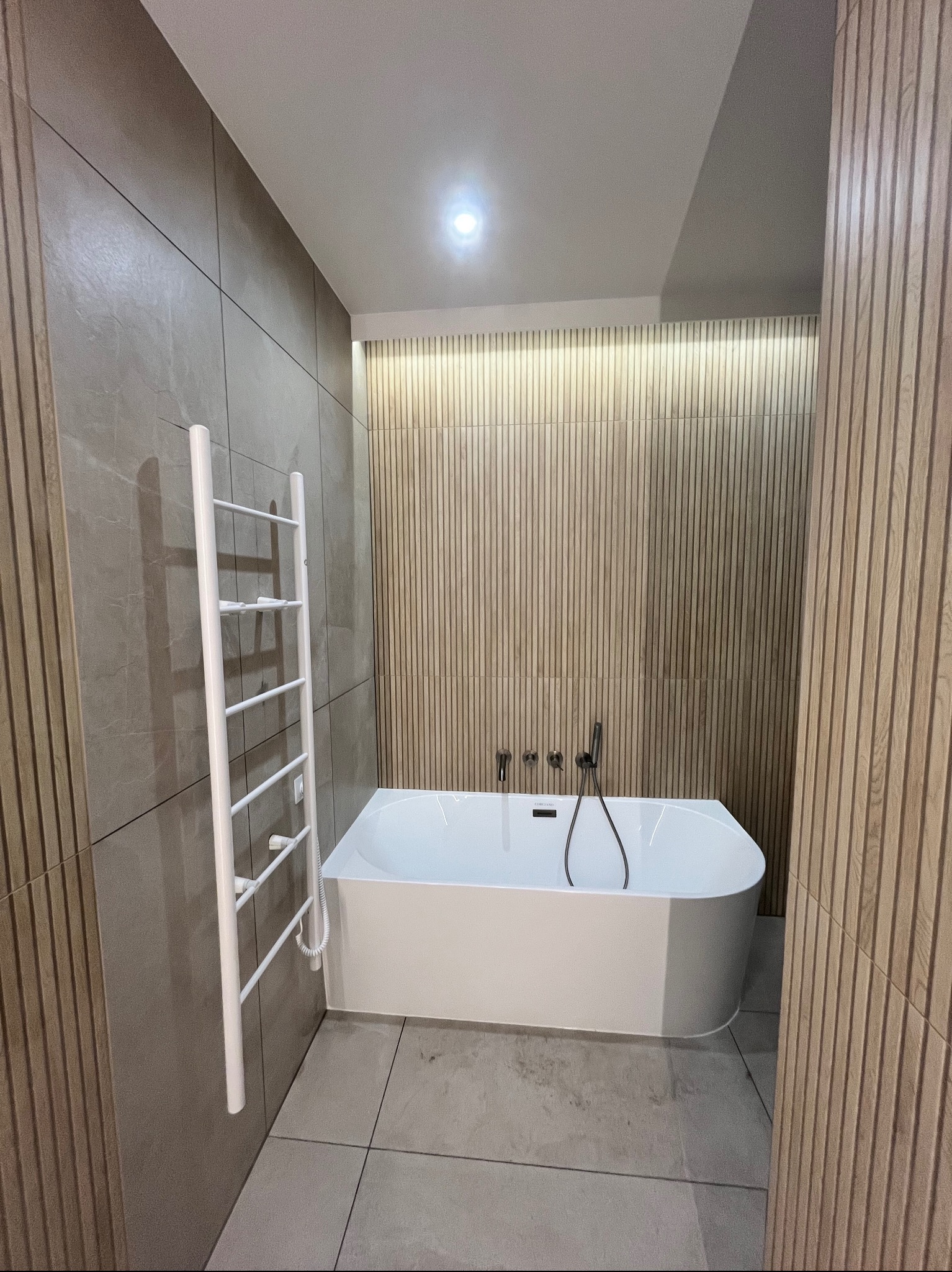 Nowoczesna łazienka z wolnostojącą wanną, grzejnikiem drabinkowym i okładziną ścienną z płytek imitujących drewno. Minimalistyczny design i stonowana kolorystyka.