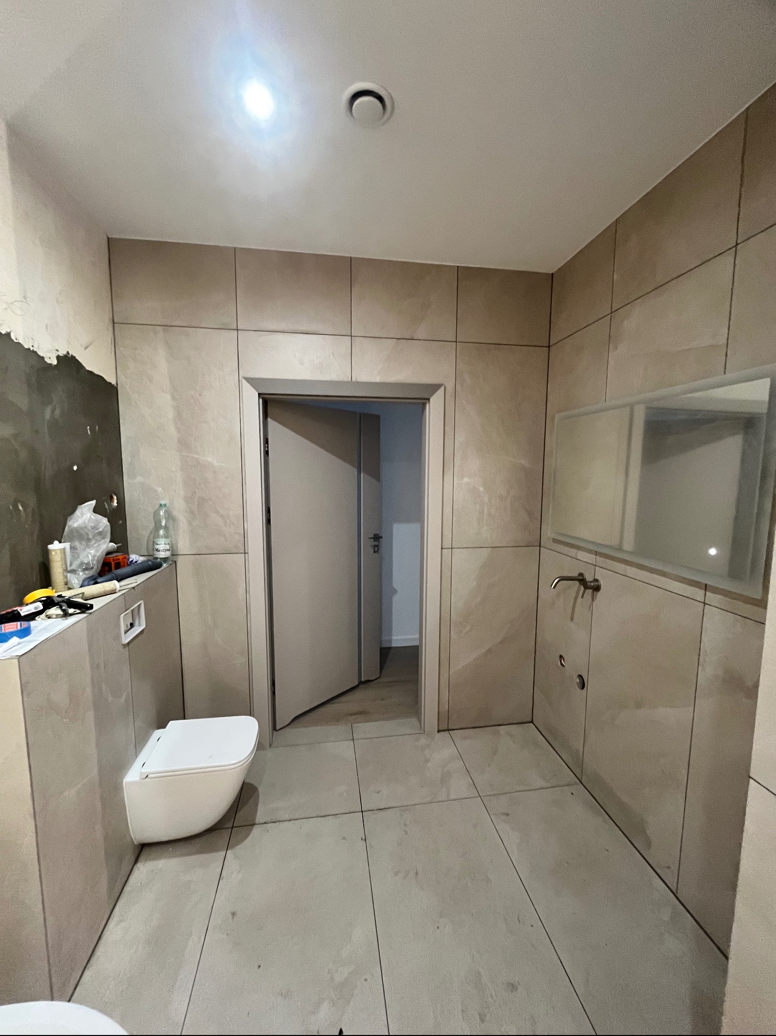 Łazienka w trakcie wykańczania: beżowe płytki imitujące marmur na ścianach i podłodze, wisząca toaleta, umywalka i lustro. Widoczne narzędzia i materiały budowlane.