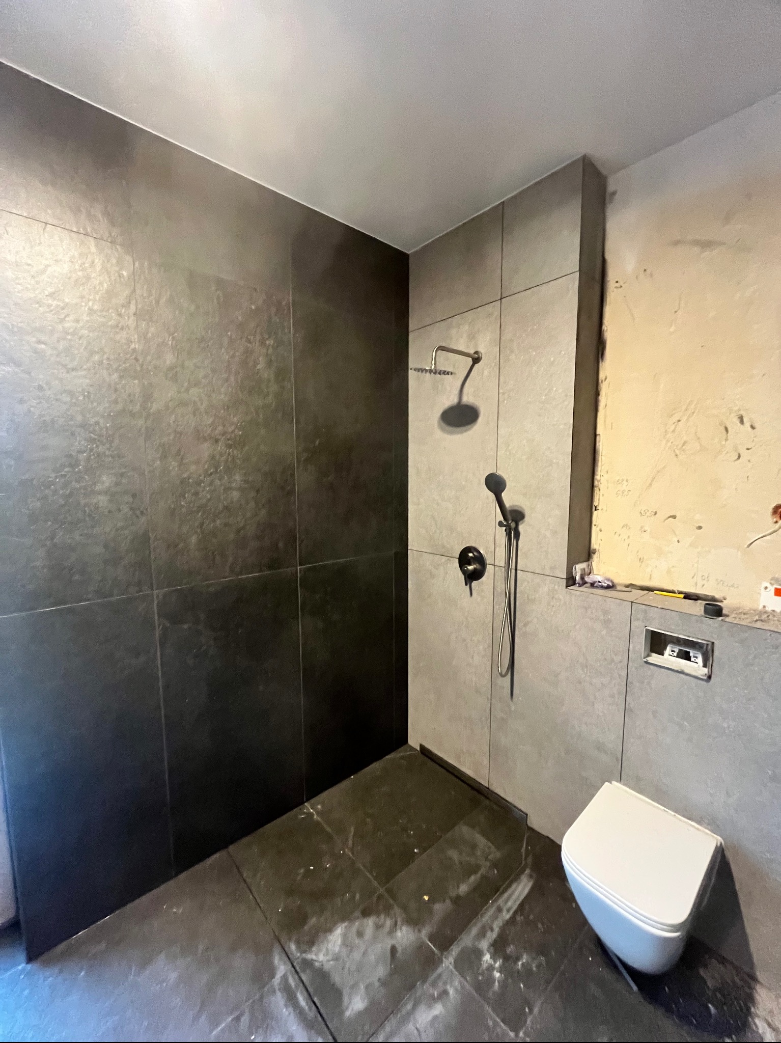 Nowoczesna łazienka w trakcie remontu: ciemne płytki podłogowe i ścienne, zamontowana toaleta, widoczna instalacja prysznicowa i fragment ściany przed wykończeniem.
