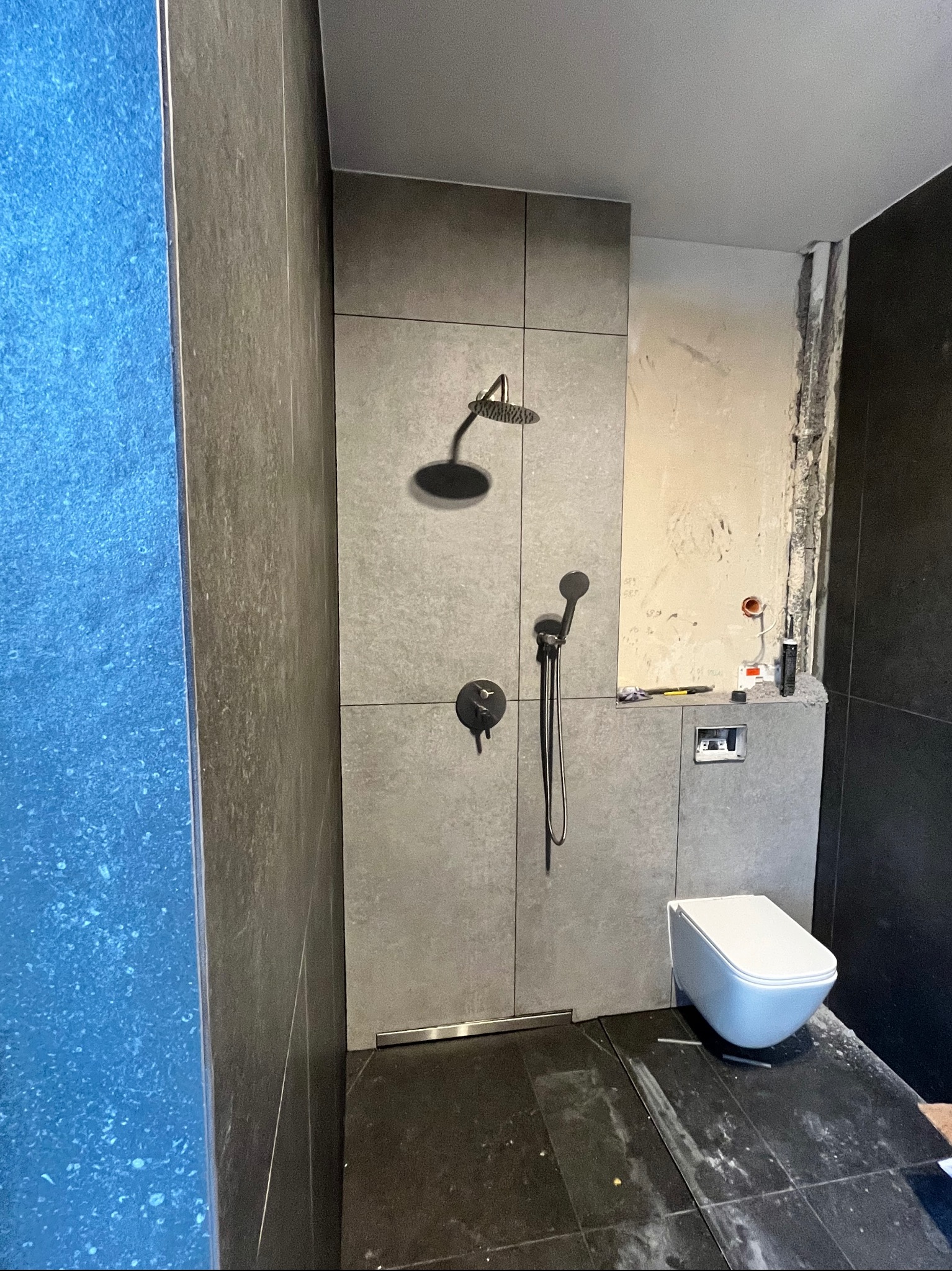 Szara łazienka w trakcie remontu: widoczna kabina prysznicowa z czarną armaturą, wisząca toaleta i fragment ściany przed wykończeniem.