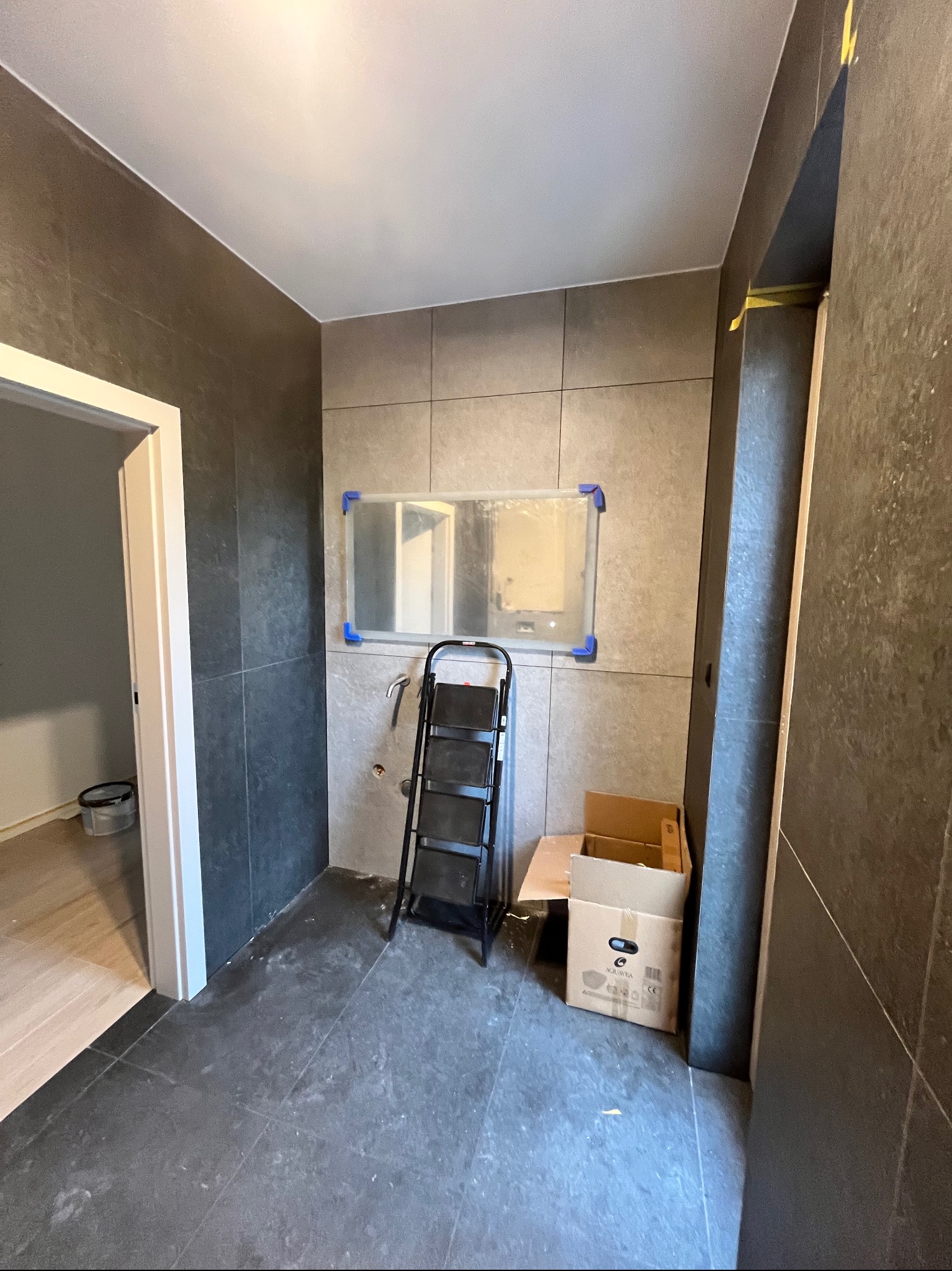 Szare płytki na ścianach i podłodze w łazience w trakcie remontu. Widoczna drabina, kartony i zabezpieczone lustro.