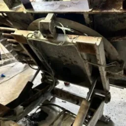 Metalowa maszyna przemysłowa z hydraulicznymi cylindrami, widoczne ślady korozji i zużycia, wnętrze hali produkcyjnej w tle.