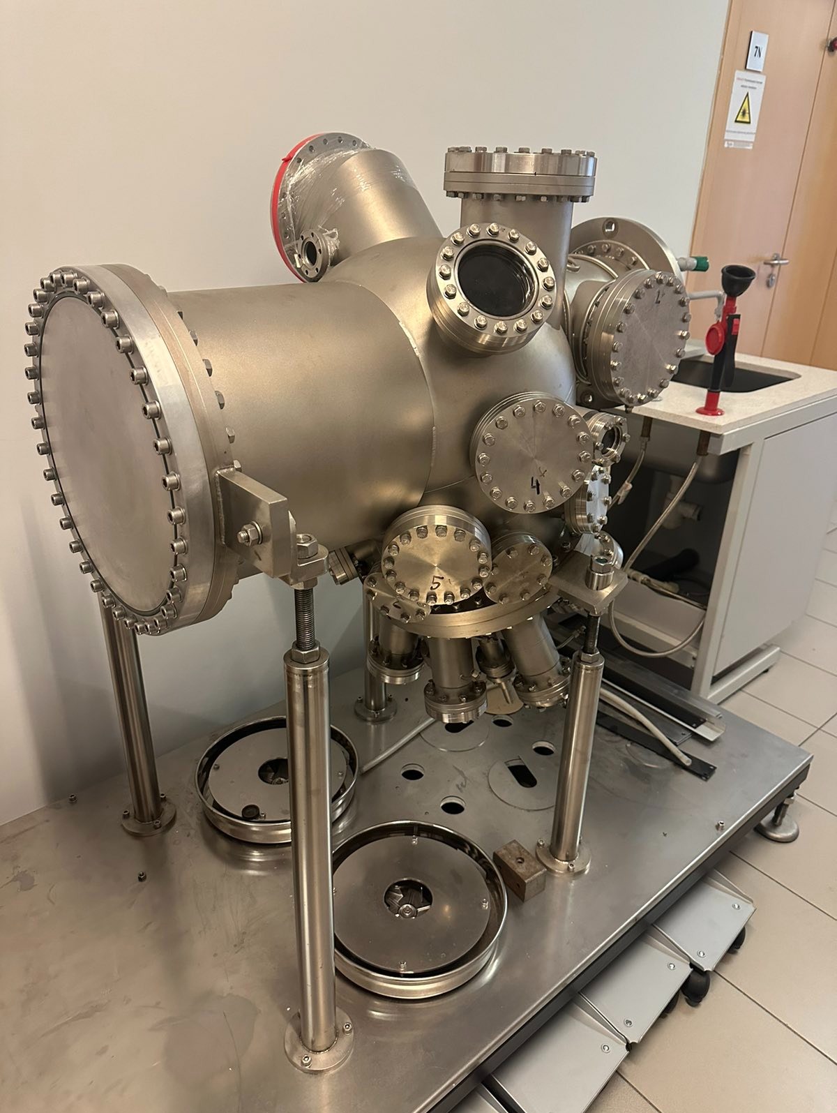 Metalowa komora próżniowa na regulowanych nogach, z licznymi przyłączami i okienkami obserwacyjnymi, umieszczona w laboratorium. Widoczny zlew i ostrzeżenie.