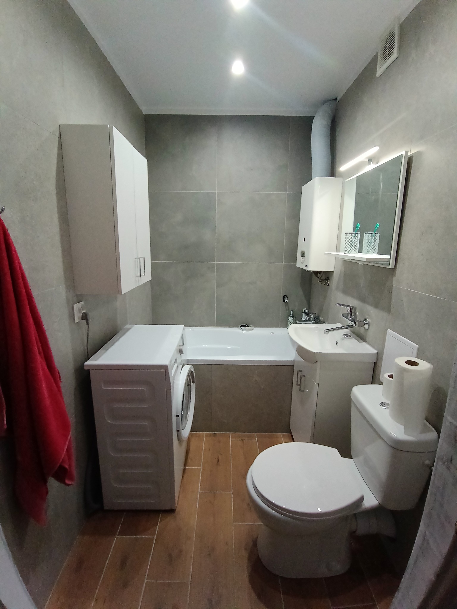 Wykończona łazienka w szarościach z brązową podłogą. Widoczna pralka, toaleta, umywalka z szafką, wanna i szafka wisząca. Czerwony ręcznik dodaje koloru.