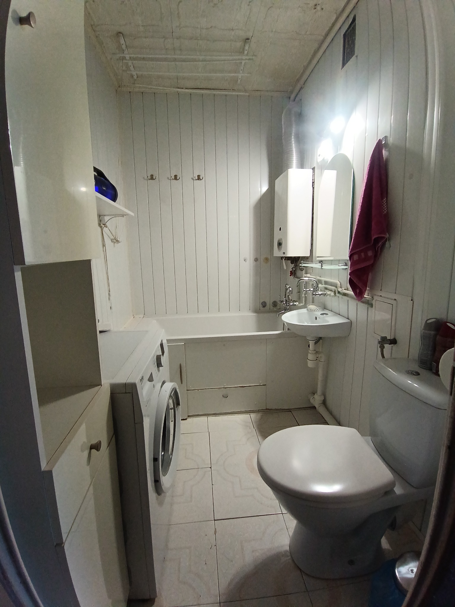 Biała łazienka z wanną, umywalką, toaletą i pralką. Ściany wyłożone panelami, widoczne rury i bojler. Ujęcie z perspektywy wejścia, oświetlenie górne.