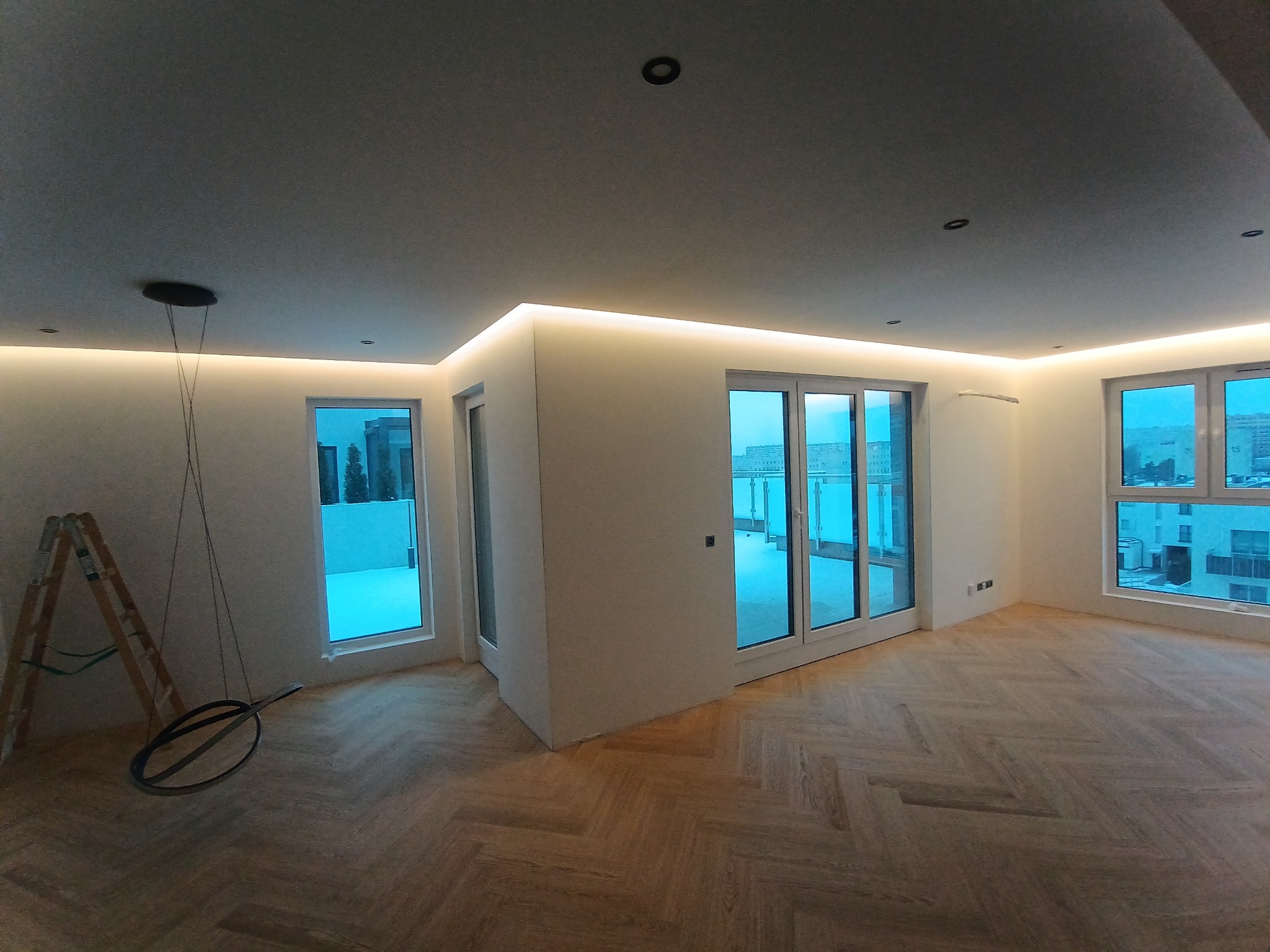 Nowoczesne wnętrze z parkietem w jodełkę, oświetleniem LED podsufitowym i dużymi oknami balkonowymi. Widoczna drabina i lampa wisząca.