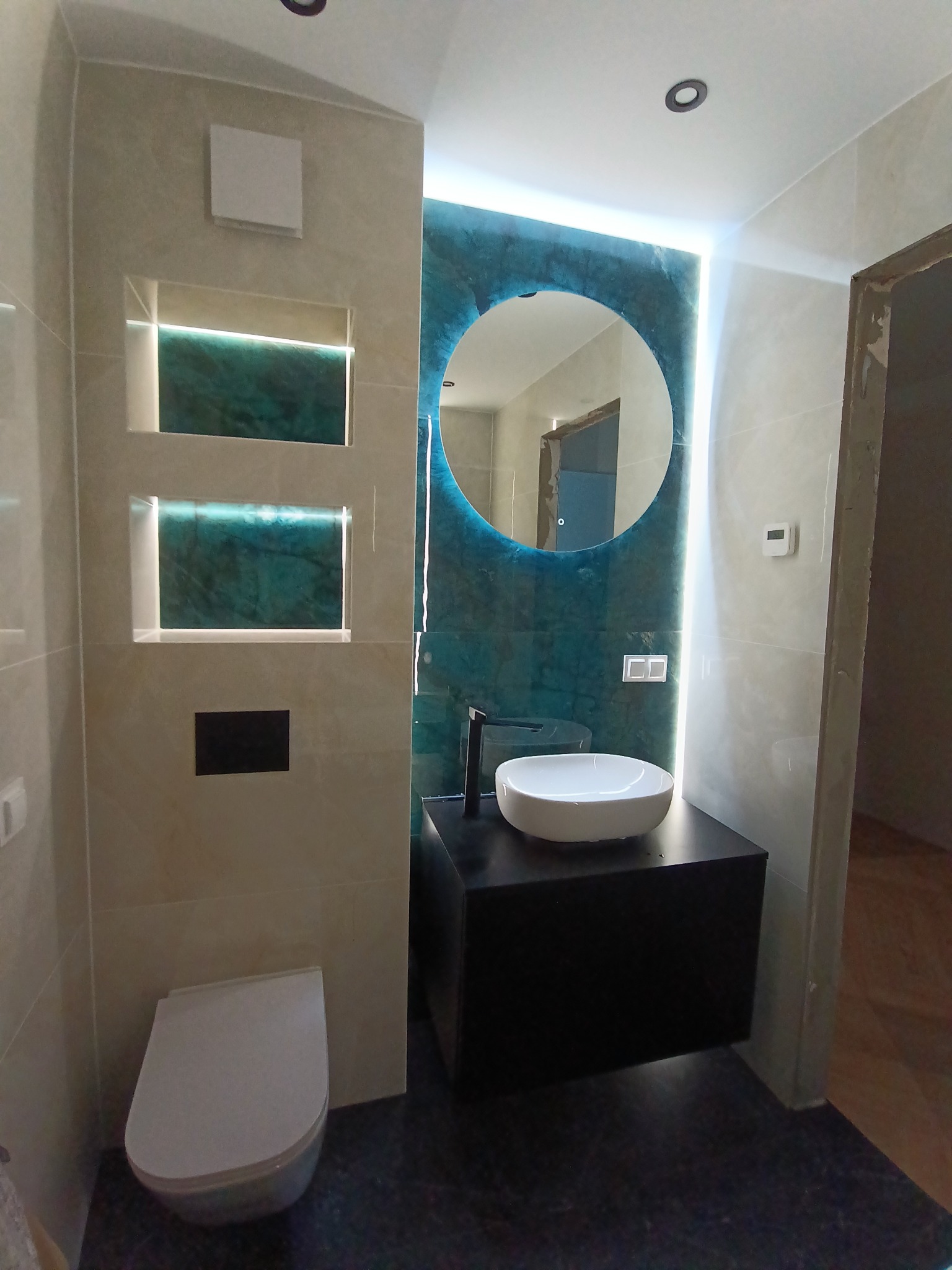 Nowoczesna łazienka z podświetlanym lustrem i wnękami, beżowe płytki, wisząca toaleta i czarna szafka pod umywalką. Minimalistyczny design z akcentem turkusowej ściany.