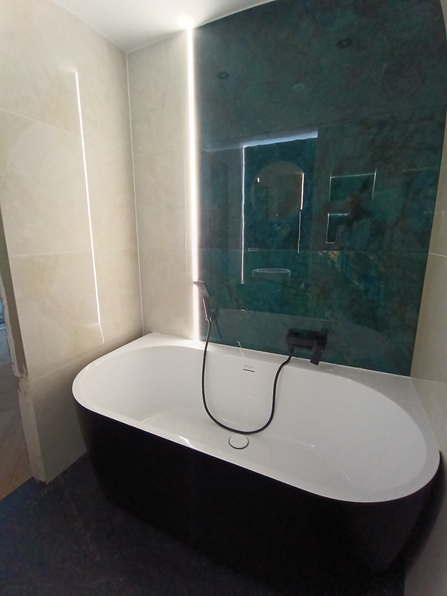 Nowoczesna łazienka z czarną wanną, podświetlanymi panelami ściennymi i zielonymi płytkami imitującymi marmur. Minimalistyczny design, eleganckie wykończenie.