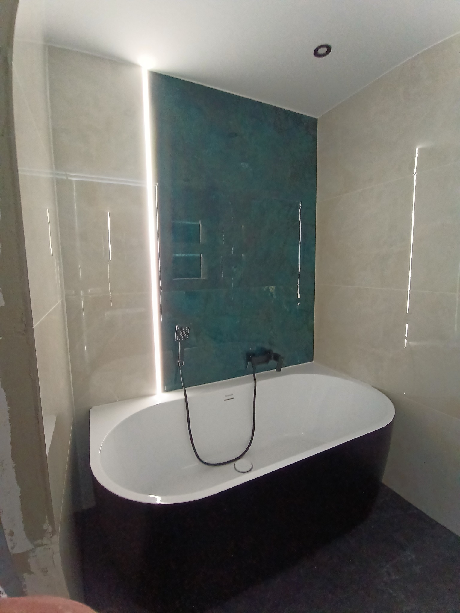 Nowoczesna łazienka z czarną wanną, zieloną ścianą i subtelnym oświetleniem LED. Minimalistyczny design, jasne płytki i ciemna podłoga tworzą elegancki kontrast.