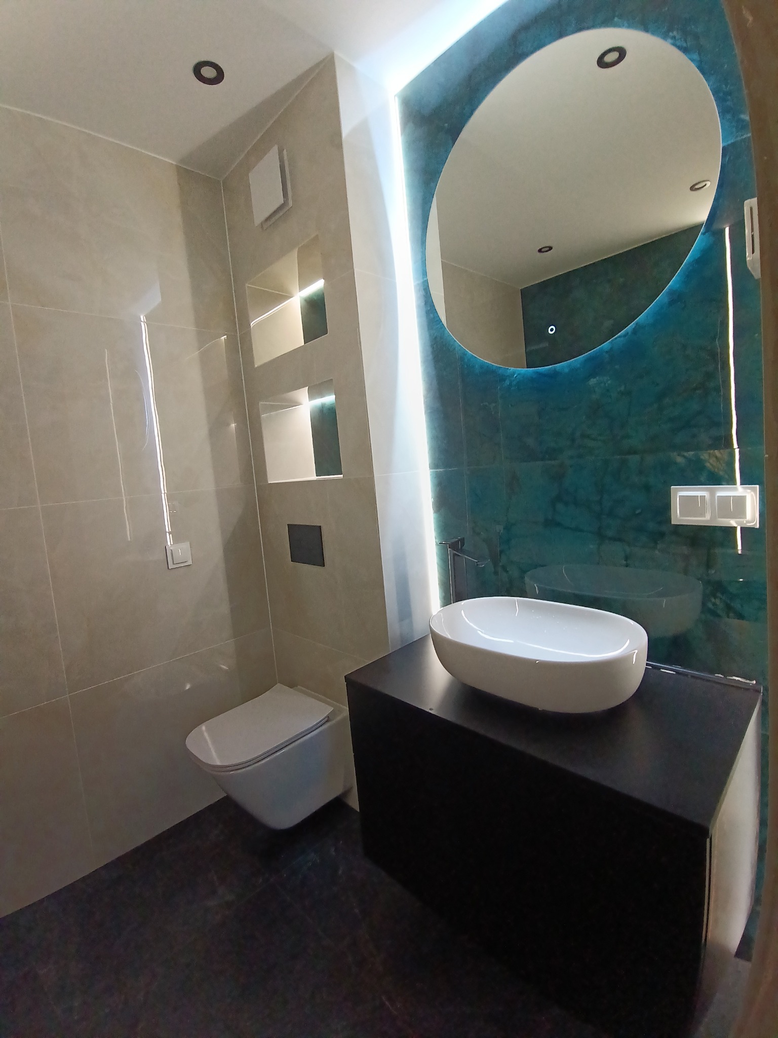 Nowoczesna łazienka z owalnym lustrem podświetlonym LED, wiszącą toaletą i umywalką nablatową na czarnej szafce. Beżowe płytki i akcent turkusowej ściany.