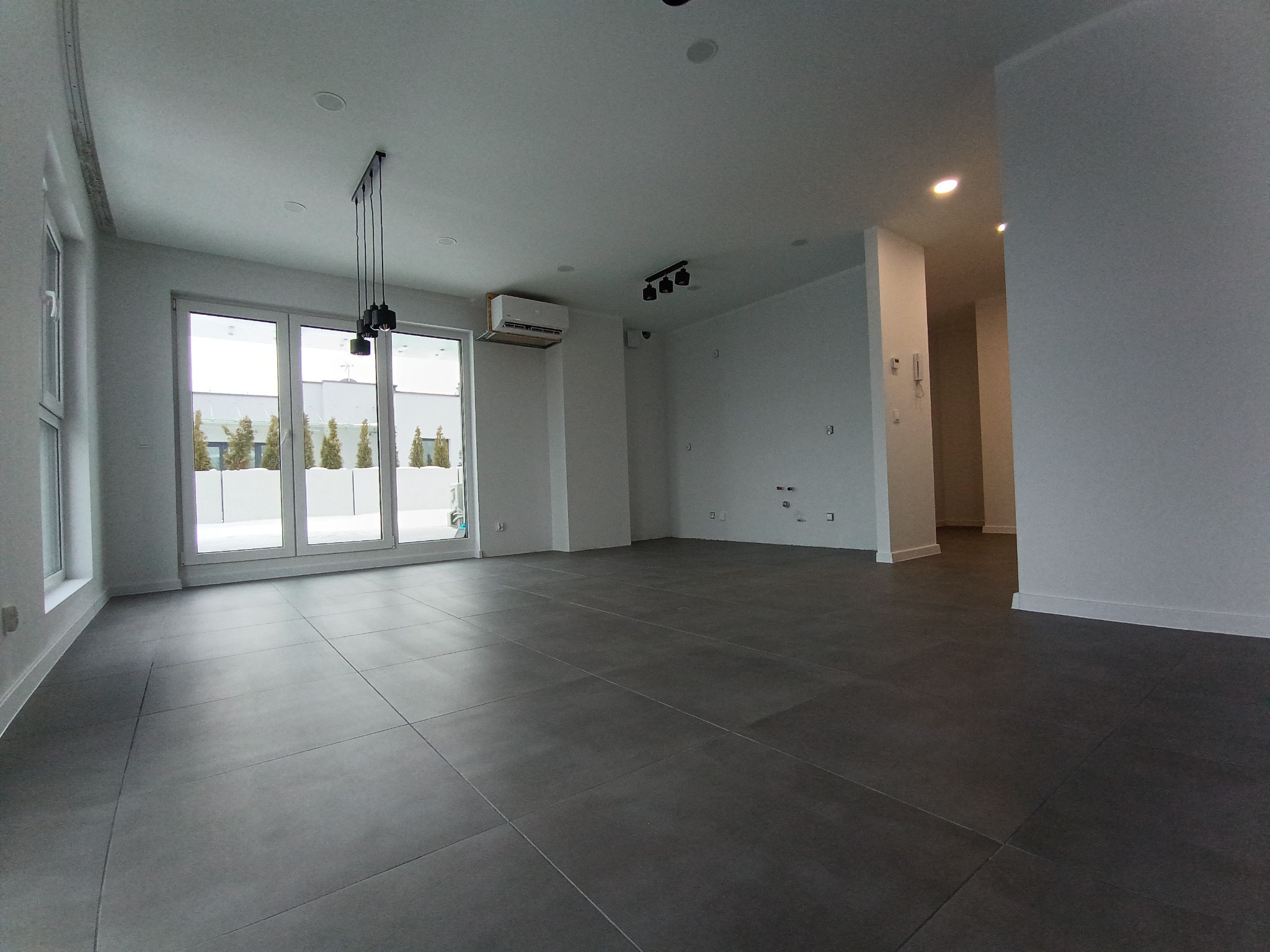 Nowoczesne wykończenie wnętrza w Jastrzębiu-Zdroju: szare płytki podłogowe, białe ściany, duże okna balkonowe z widokiem na zewnątrz i minimalistyczne oświetlenie.