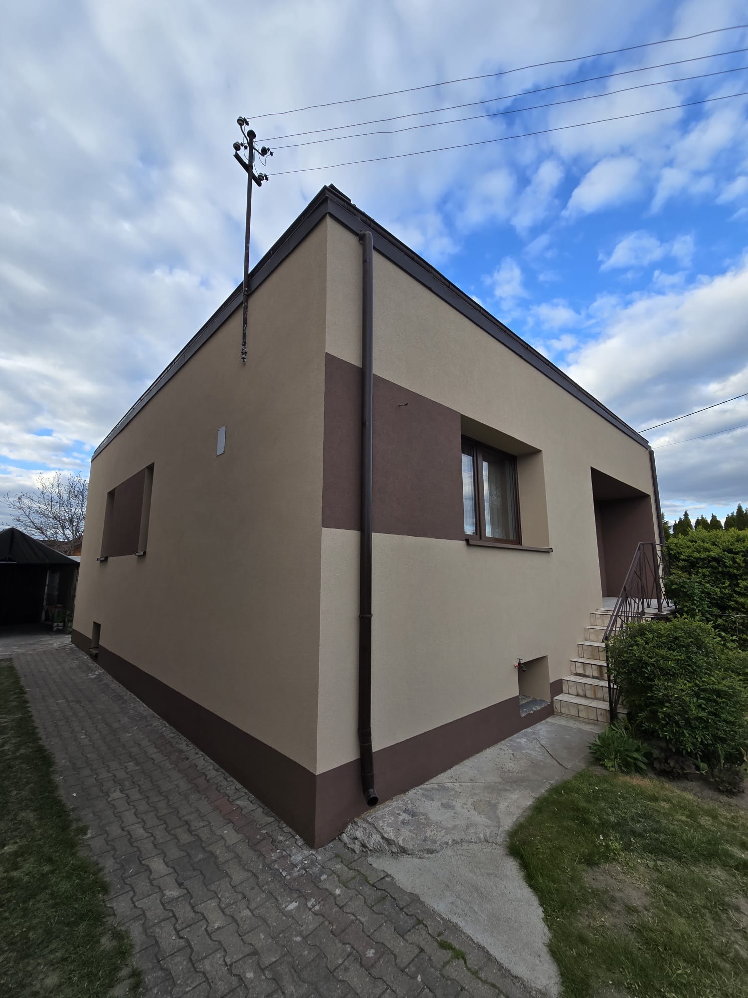 Dwukolorowa elewacja budynku z brązowymi akcentami, widok z boku z kostką brukową i zielenią. Słup elektryczny przy budynku na tle pochmurnego nieba.