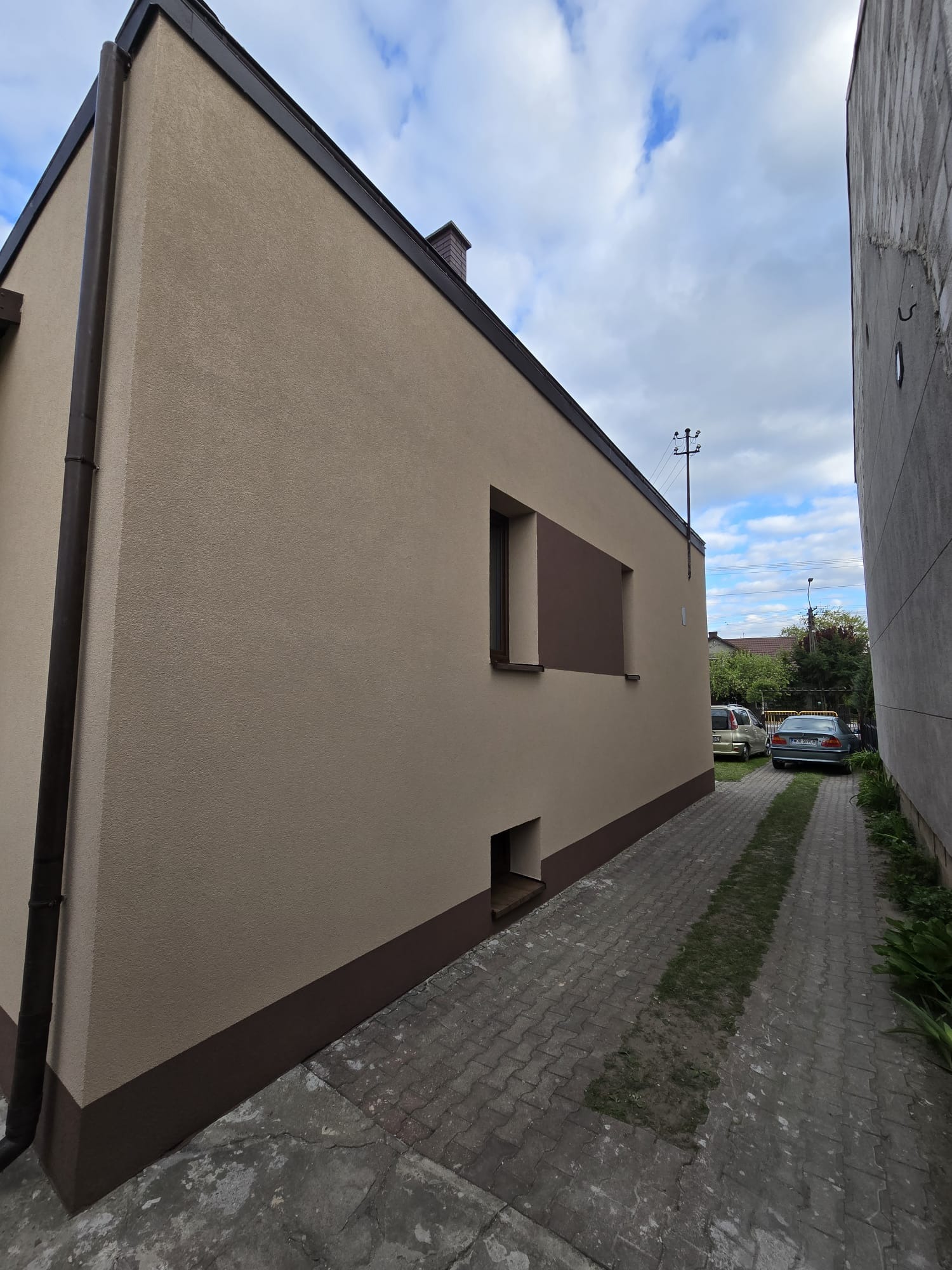 Beżowa elewacja domu z brązowymi akcentami, widok z boku na tle pochmurnego nieba i brukowanej drogi dojazdowej.