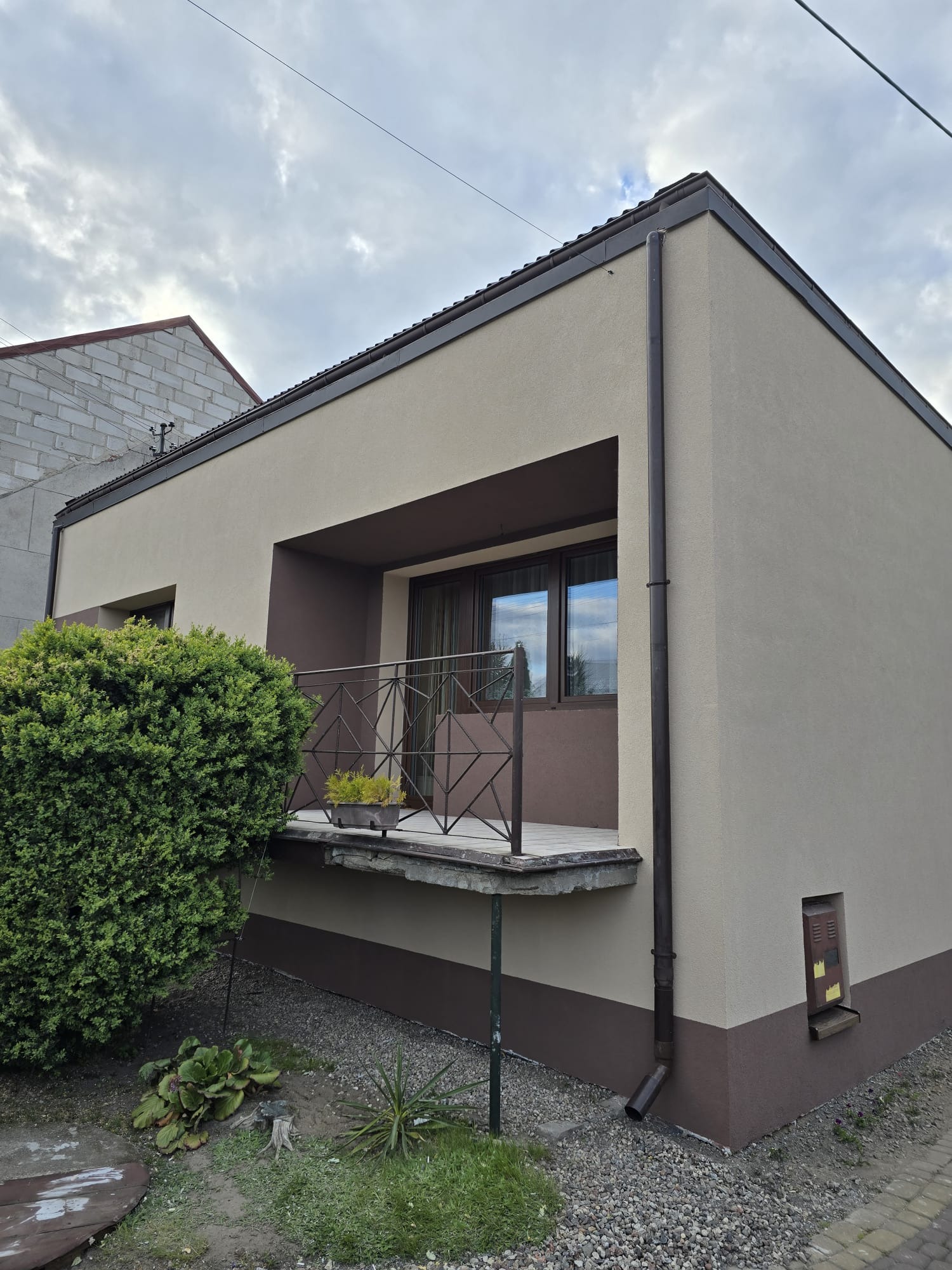 Dwukolorowa elewacja domu z balkonem, widok z boku. Brązowe detale architektoniczne i roślinność w otoczeniu budynku. Nowoczesna estetyka budynku.