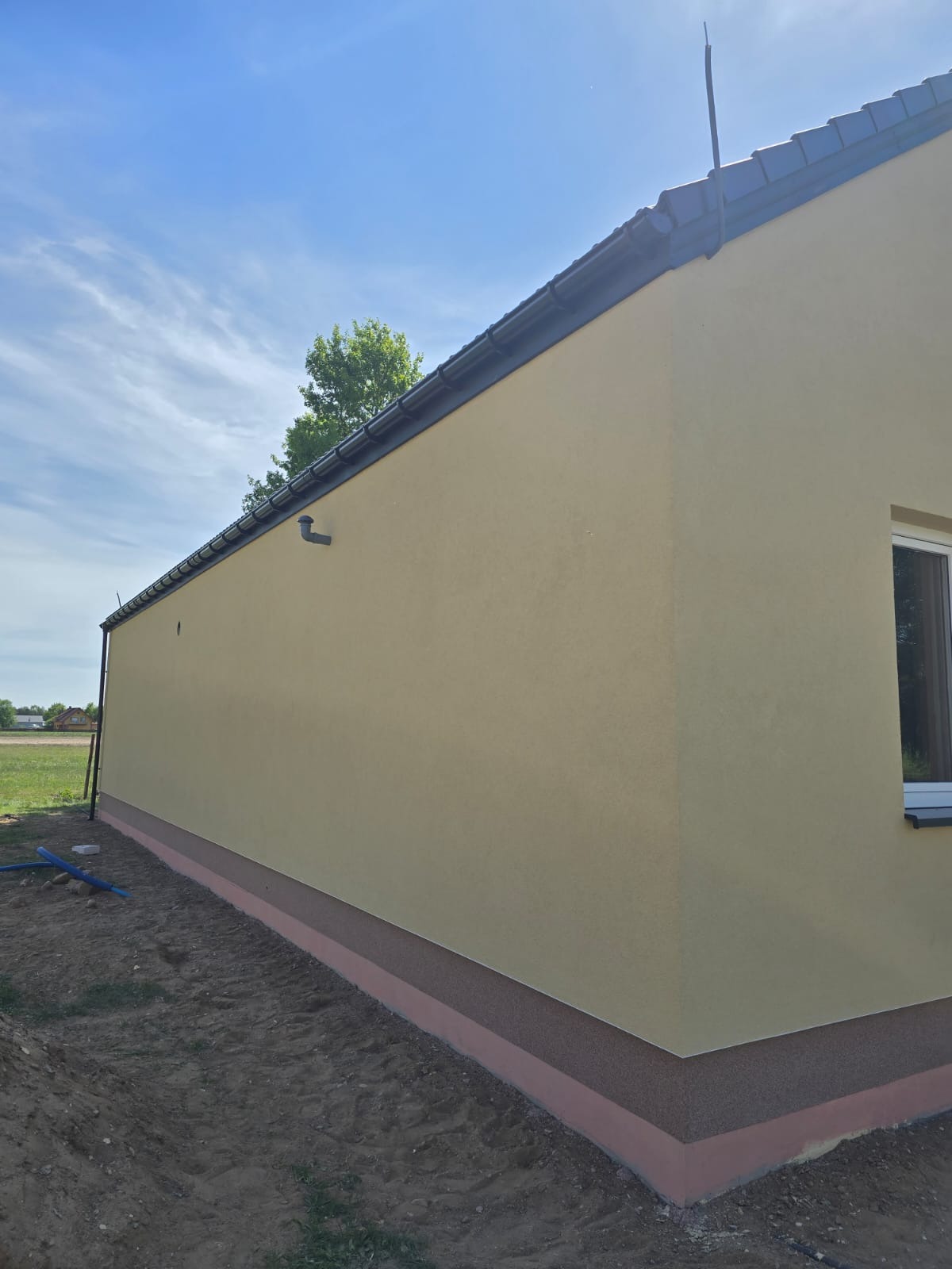 Ściana budynku po nałożeniu tynku strukturalnego w odcieniach beżu, z widocznym oknem i ciemnym dachem, na tle błękitnego nieba i zieleni.