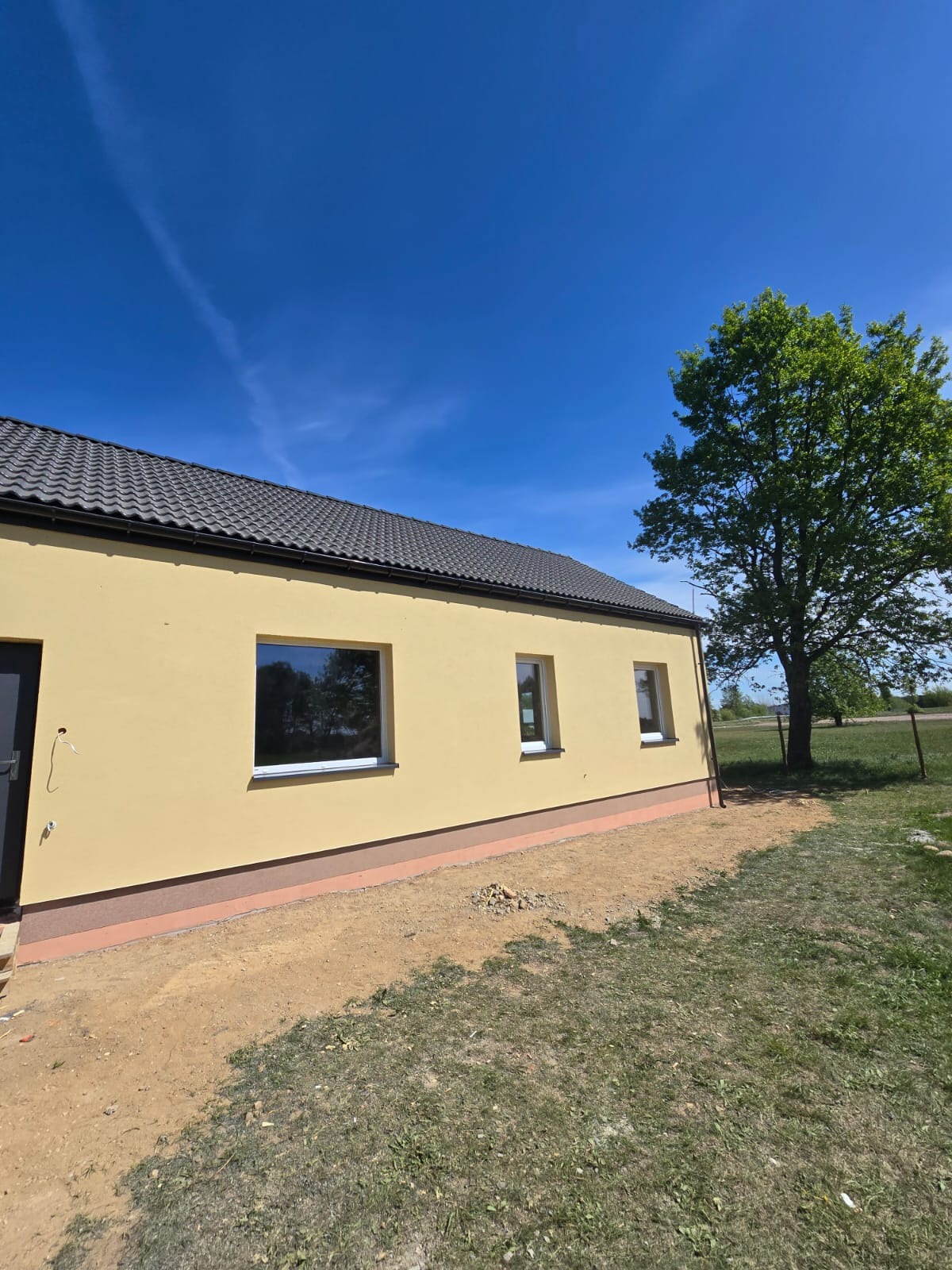 Żółta elewacja domu jednorodzinnego z szarą dachówką i białymi oknami w słoneczny dzień, widok z boku na tle zieleni i błękitnego nieba.