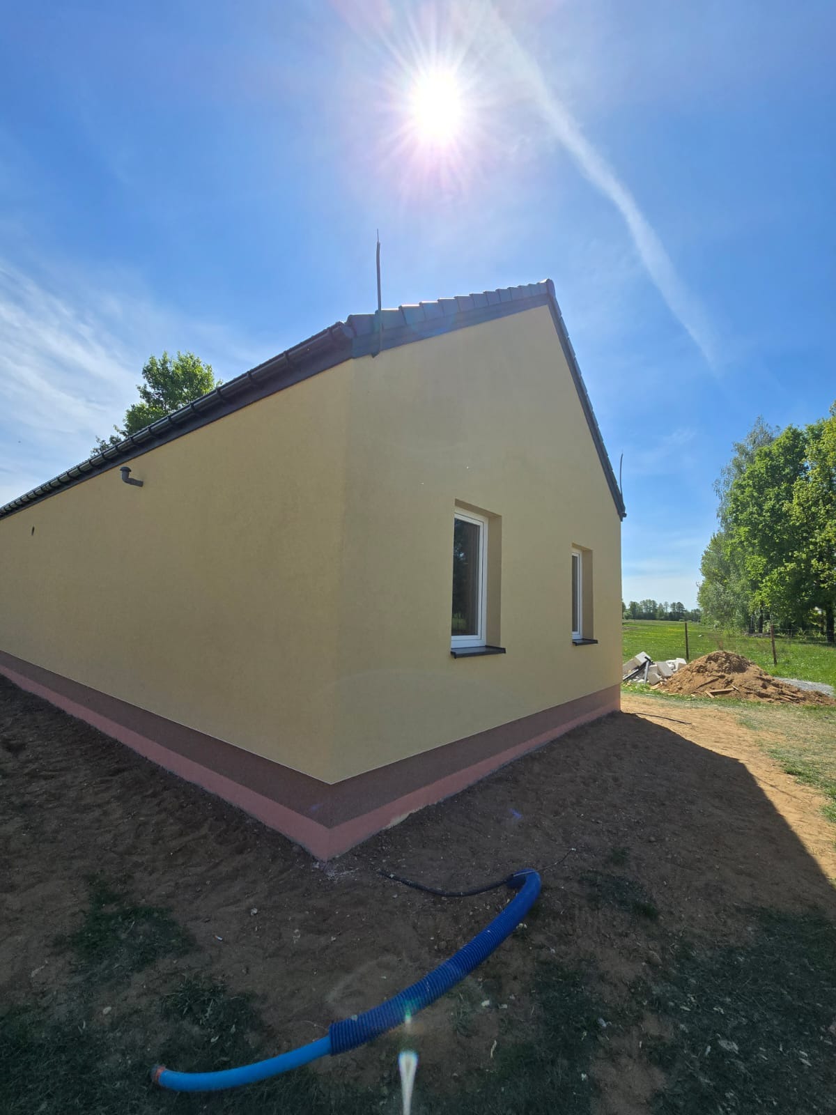 Żółty dom jednorodzinny z nową elewacją, widok z boku w słoneczny dzień. Ciemna podmurówka i białe ramy okienne. W tle zieleń i błękitne niebo.