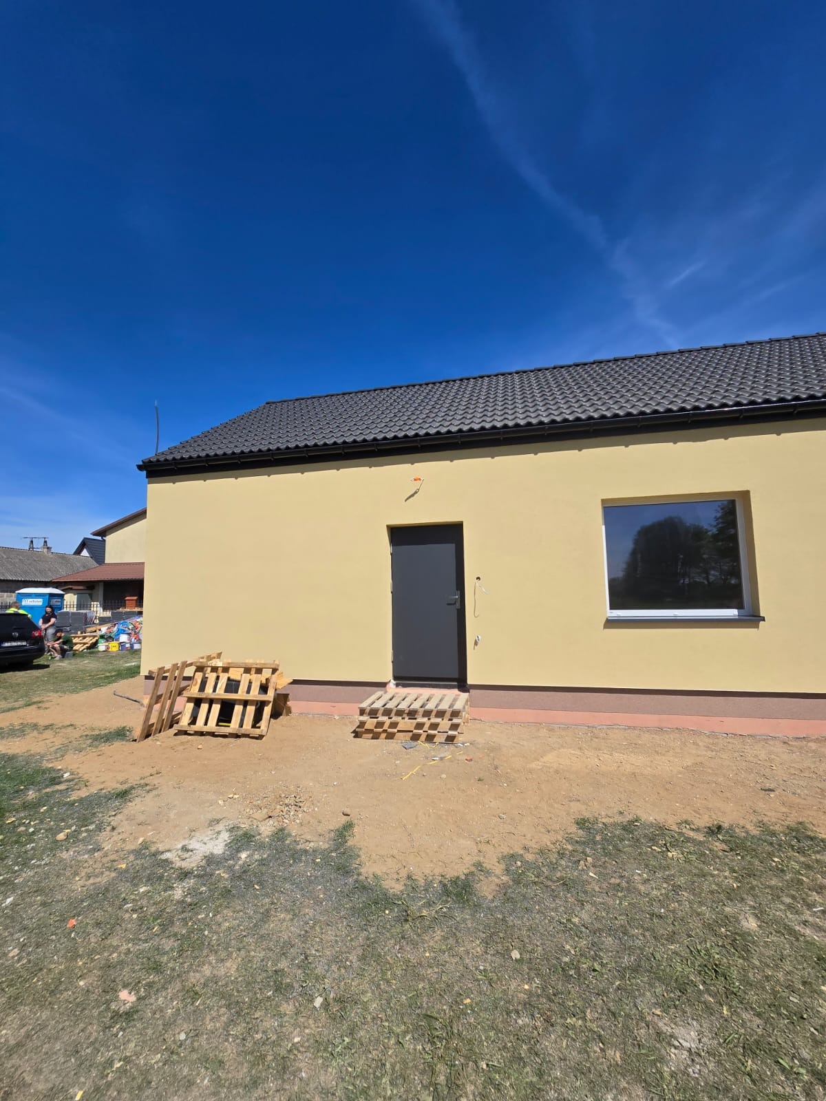 Jasnożółty budynek z szarą stolarką i dachem. Widoczne palety budowlane. Świeżo wykonana elewacja w słoneczny dzień. Ciemne okno odbija otoczenie.